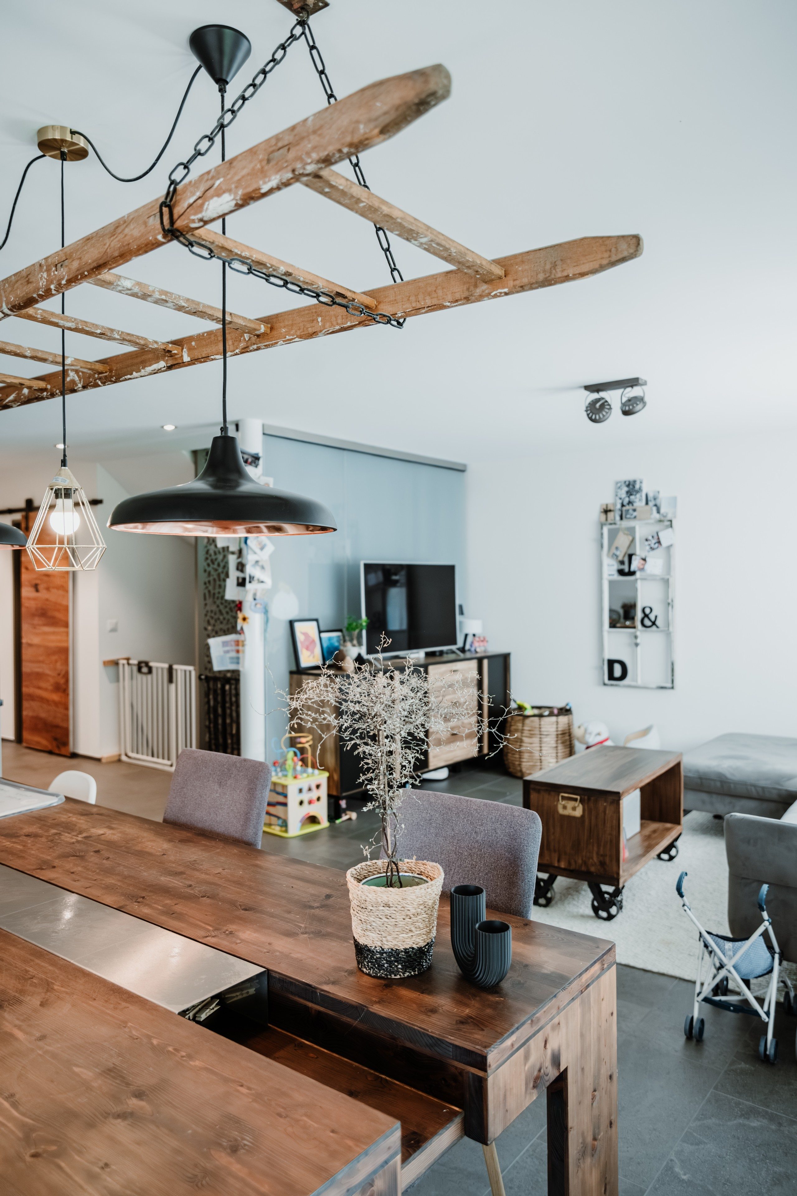 Ein Wohn- und Essbereich in einem modernen Einfamilienhaus. Vorne steht ein massiver Holztisch mit Metallelementen. Oben hängt eine Lampe, welche aus einer alten Holzleiter gebaut wurde. Hinten im Bild sieht man verschiedene Holzmöbel und Spielsachen und einen TV.