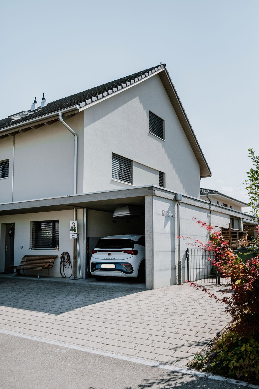 Modernes zweistöckiges Wohnhaus mit Carport, unter dem ein weisses Auto der Marke Cupra geparkt ist. Neben dem Carport stehen eine Holzbank und Gartengeräte. Ein Schild mit der Aufschrift «Waldhof-Allee» ist an der Hauswand angebracht. Im Vordergrund ist ein gepflasterter Gehweg und ein Strauch zu sehen.
