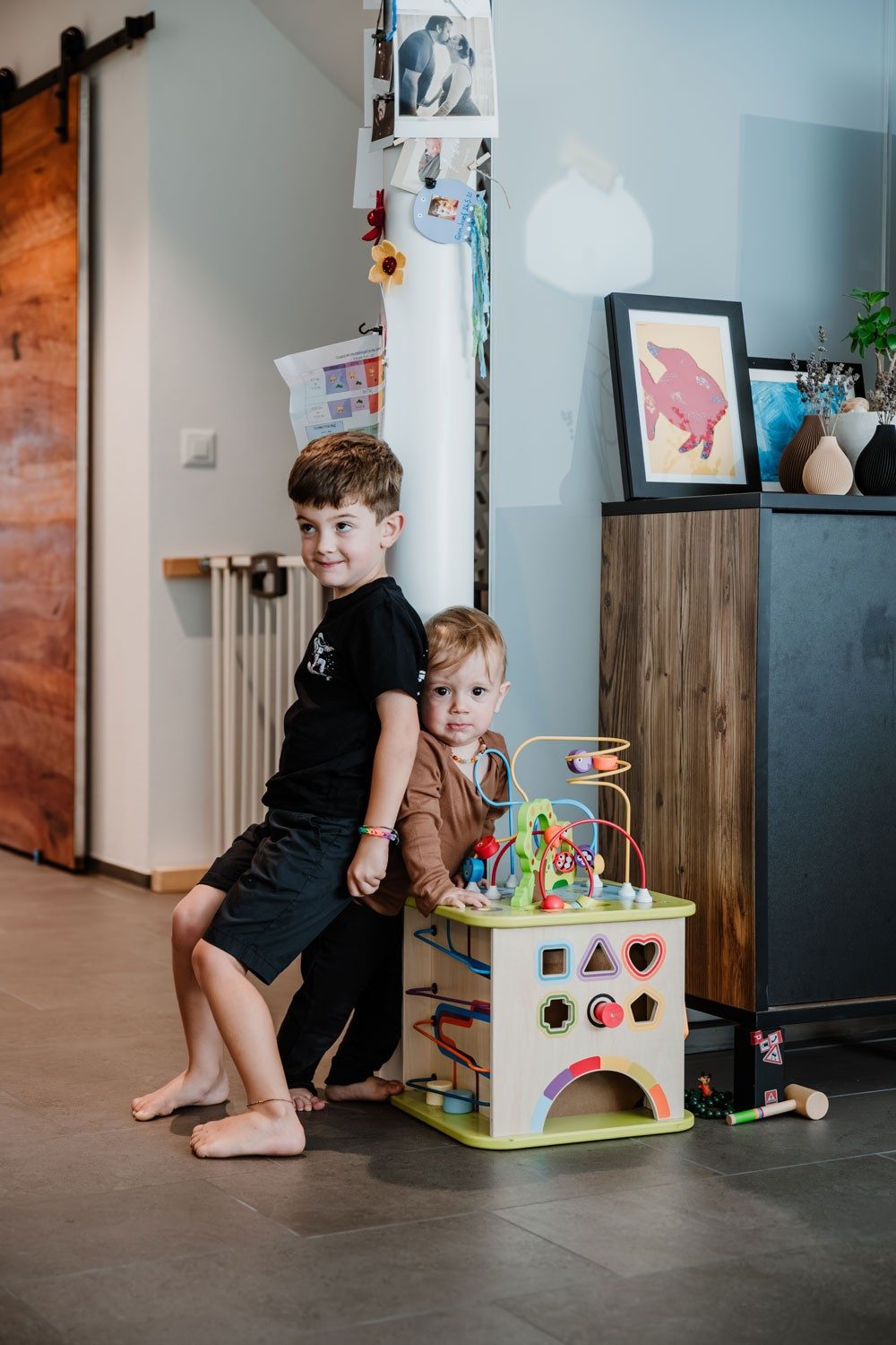 Zwei kleine Jungen stehen mit dem Rücken zueinander gelehnt und halten sich an einem Kinderspielzeug fest. Sie befinden sich in einem Wohnraum. Neben ihnen steht eine Holzkommode mit Dekoelementen. Hinter ihnen befindet sich ein Treppenkinderschutz und eine grosse Holztüre.