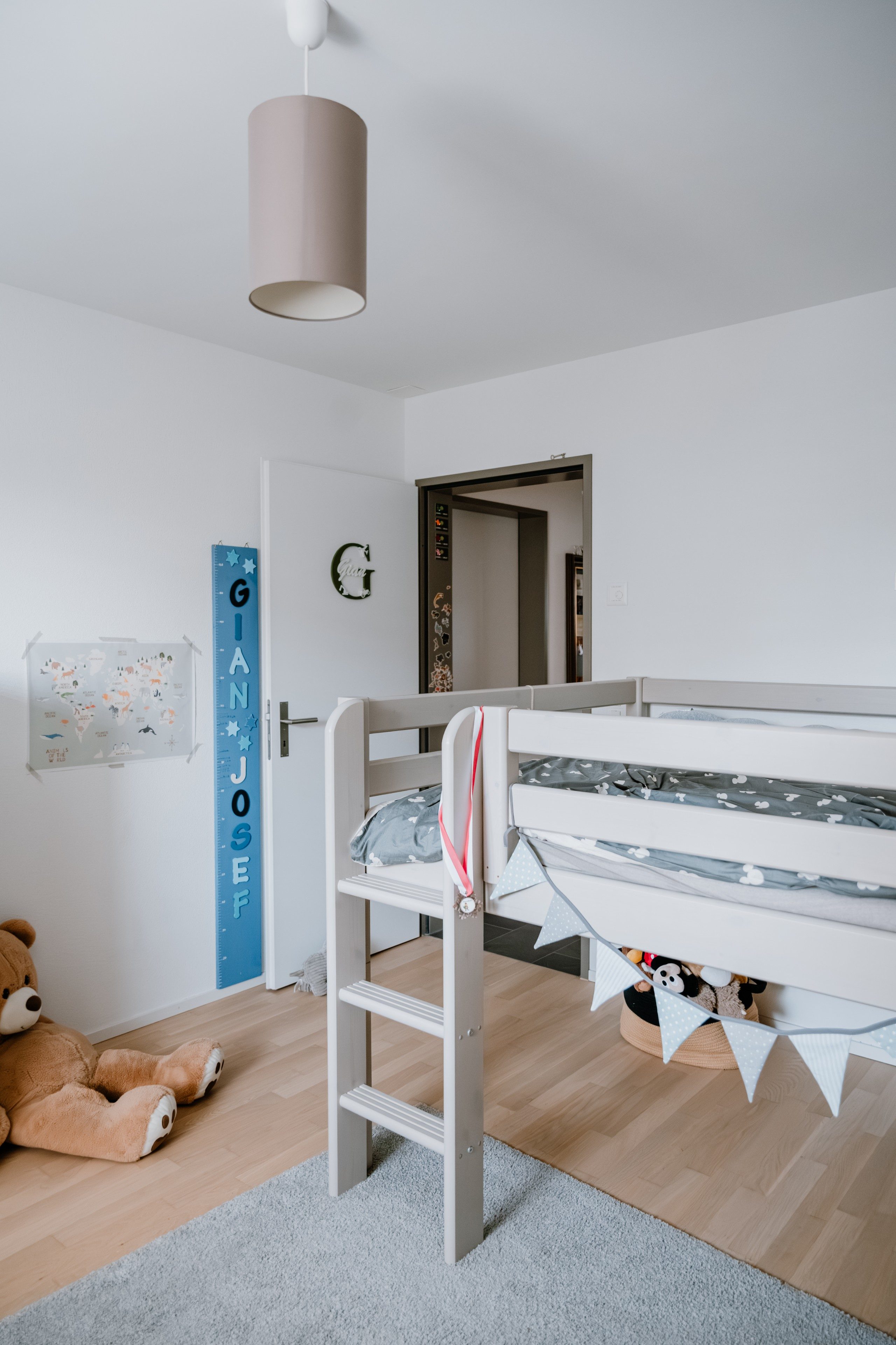 Ein Kinderzimmer mit einem weissen Kinderbett aus Holz. Es ist erhöht und über eine kleine Leiter zugänglich. Am Bett sind kleine Fähnchen als Deko angebracht. Hintendran sitzt ein Teddy-Bär auf dem Boden. Die Tür trägt den Schriftzug G und Gian in Form einer Holzdeko. Daneben ist eine Messlatte, auf der in Grossbuchstaben Gian Josef steht.