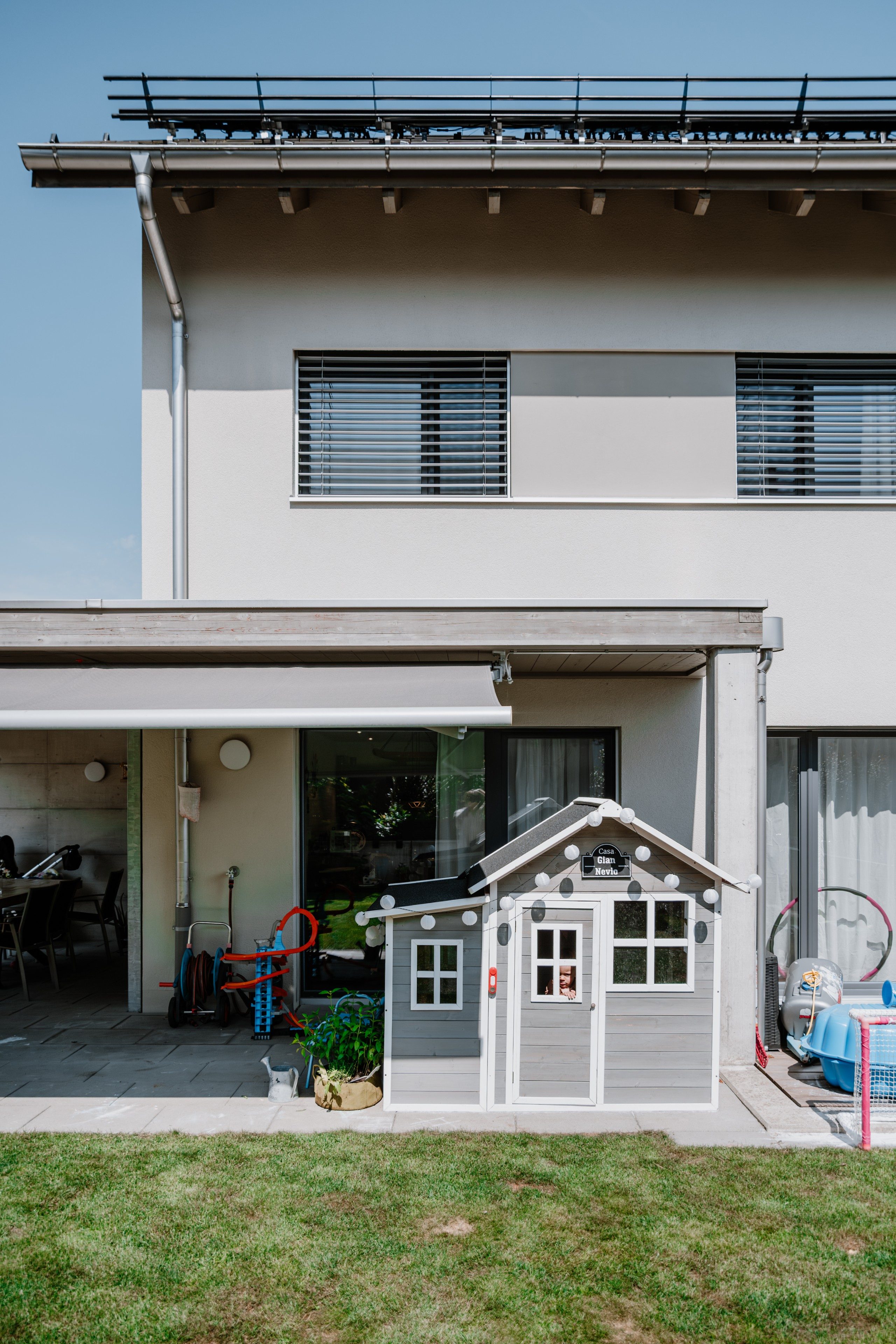 Die Aussenfassade eines modernen, zweistöckigen Hauses. Vor dem Haus steht ein Kinderspielzeughaus. Darauf befindet sich eine Tafel, auf der «Casa Gian Nevio» steht. Das kleine Haus ist mit einer Lichterkette geschmückt.