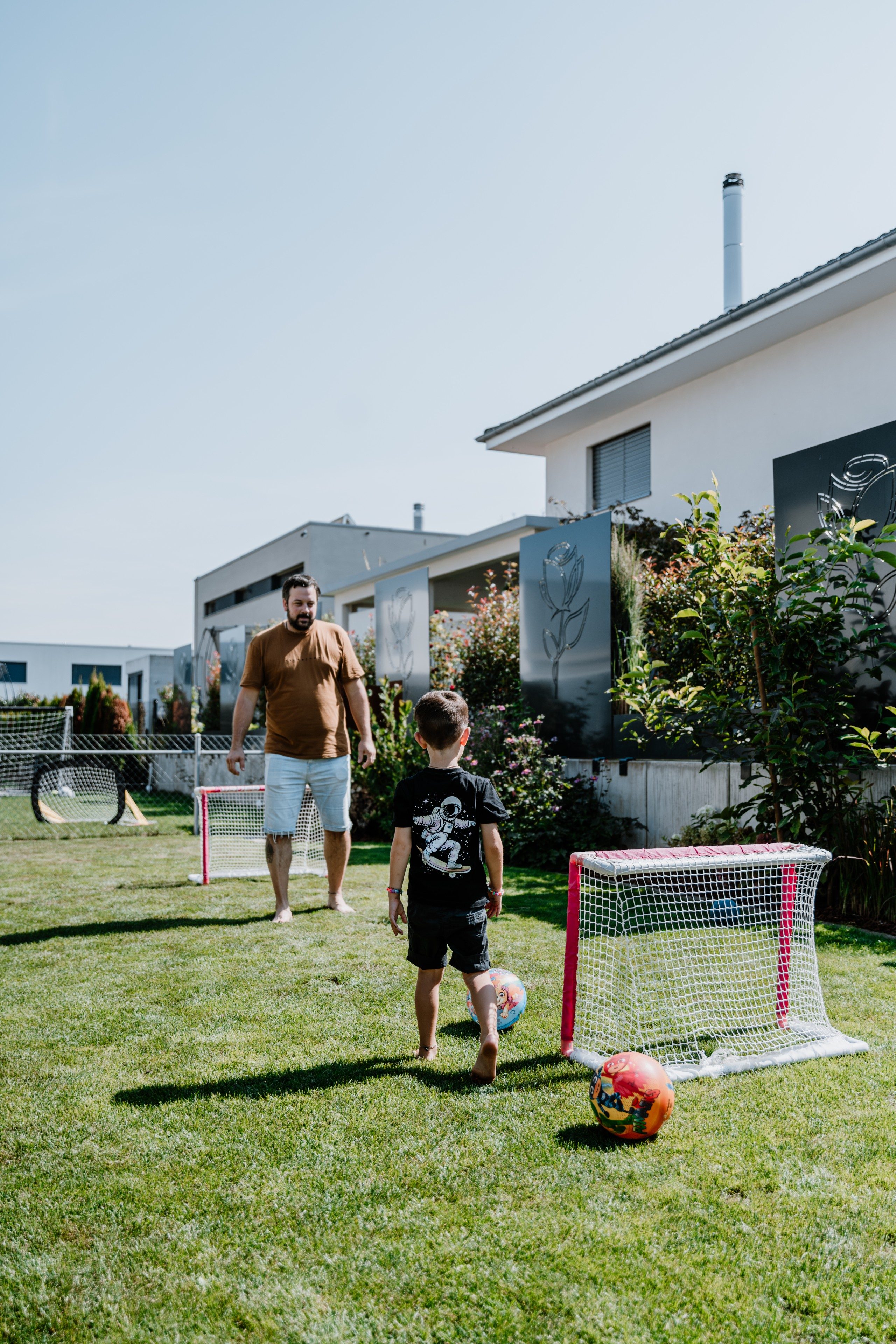 Mann mit kurzer Hose und braunem T-Shirt steht in einem Garten vor einem Kinder-Fussballtor. Vor ihm steht ein kleiner Junge mit dem Rücken zur Kamera gerichtet und ist dabei, einen Ball mit buntem Aufdruck zu schiessen. Der Junge hat auf dem T-Shirt am Rücken den Aufdruck eines Astronauten, der Skateboard fährt.