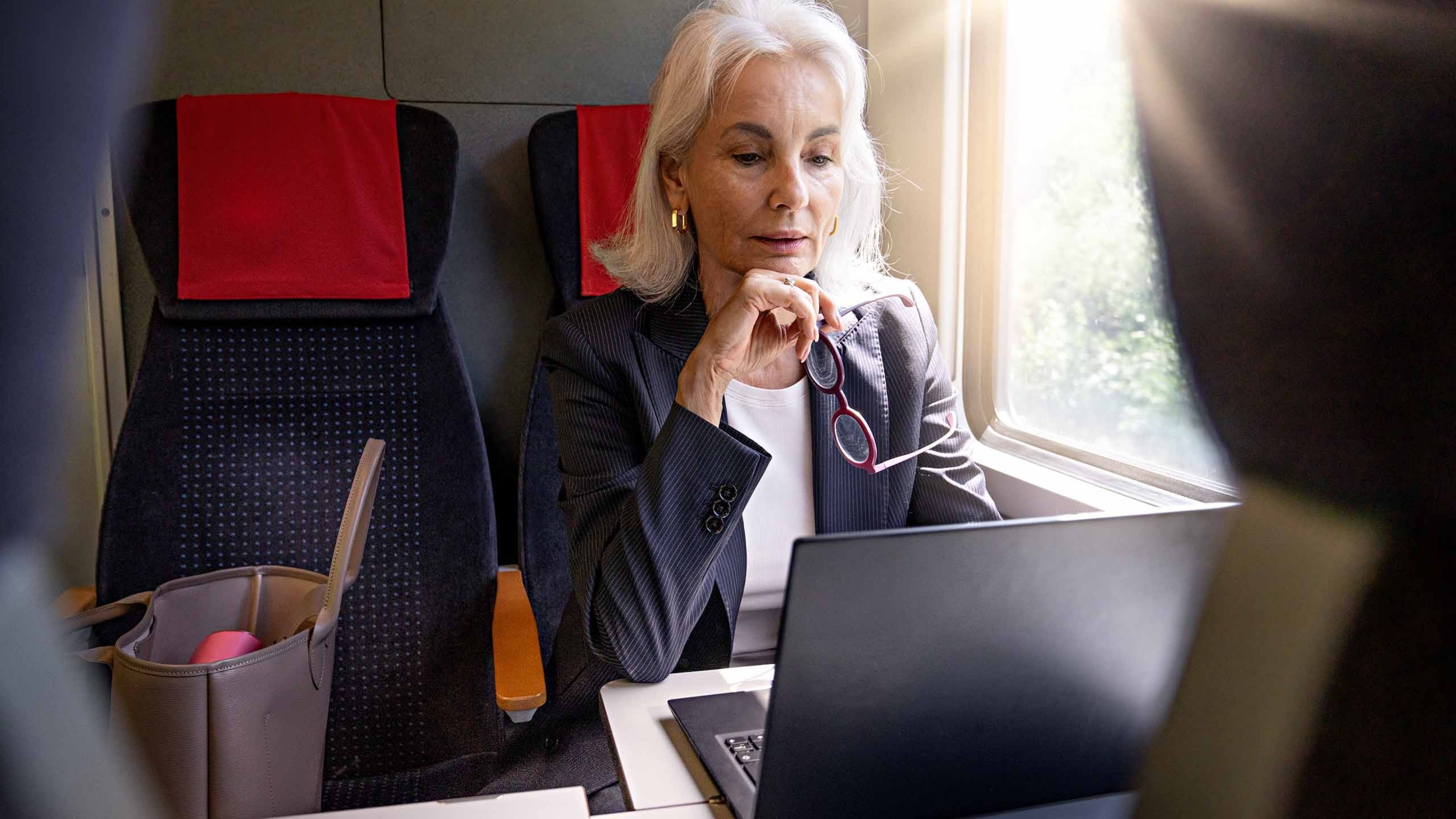 Eine Frau sitzt in einem Zugabteil und blickt auf den Bildschirm eines geöffneten Notebooks, das auf dem Tisch vor ihr steht. Neben ihr befindet sich eine offene Tasche auf dem Sitz.