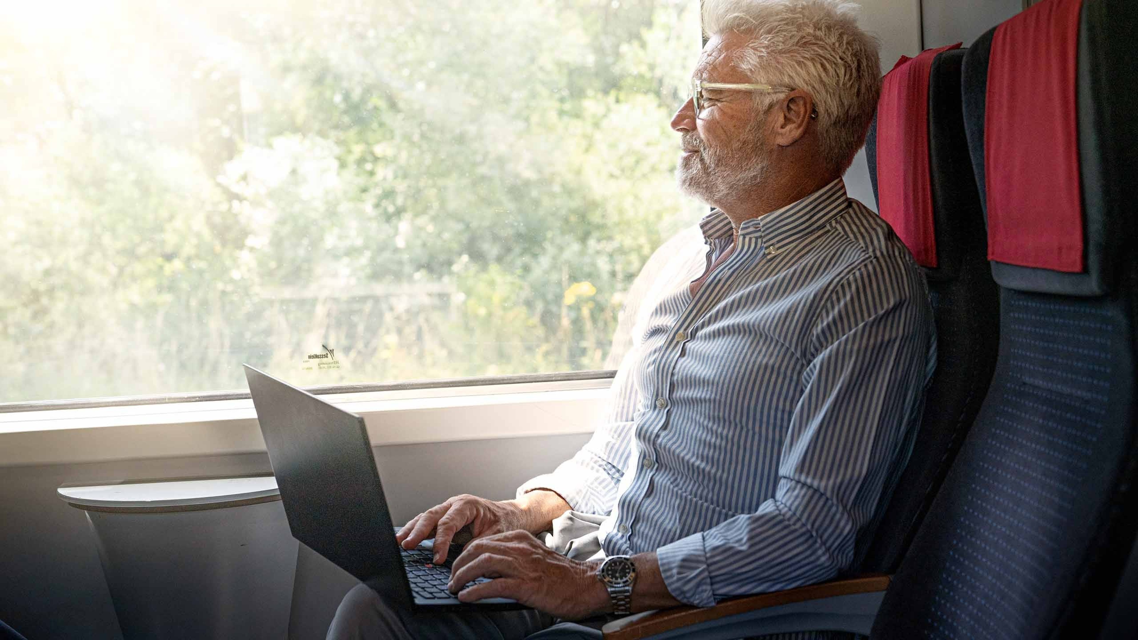 Ein Mann sitzt in einem Zugabteil mit einem geöffneten Notebook auf den Beinen. Seine Hände ruhen auf der Tastatur, während er aus dem Fenster ins Grüne blickt.