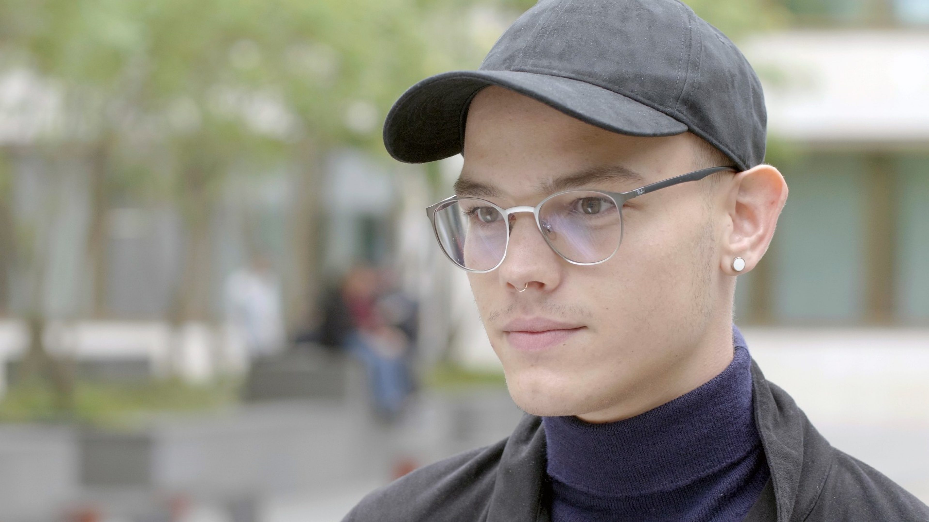 Ein Student mit Mütze und Brille steht vor einem Gebäude.
