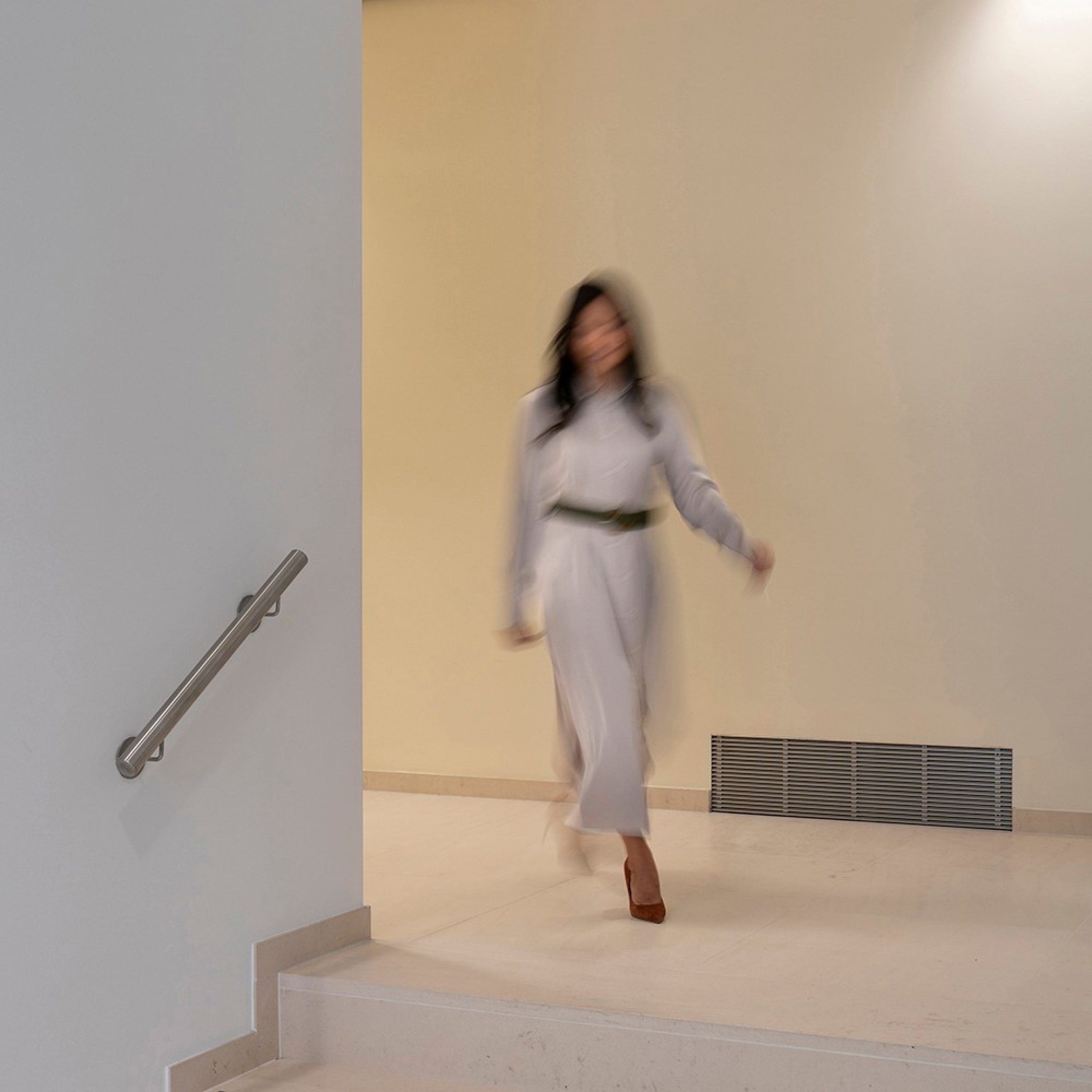 Eine elegante Frau in einem weissen Blusenkleid mit schwarzem Gürtel und braunen High Heels läuft um eine Ecke eine Treppe hinunter.