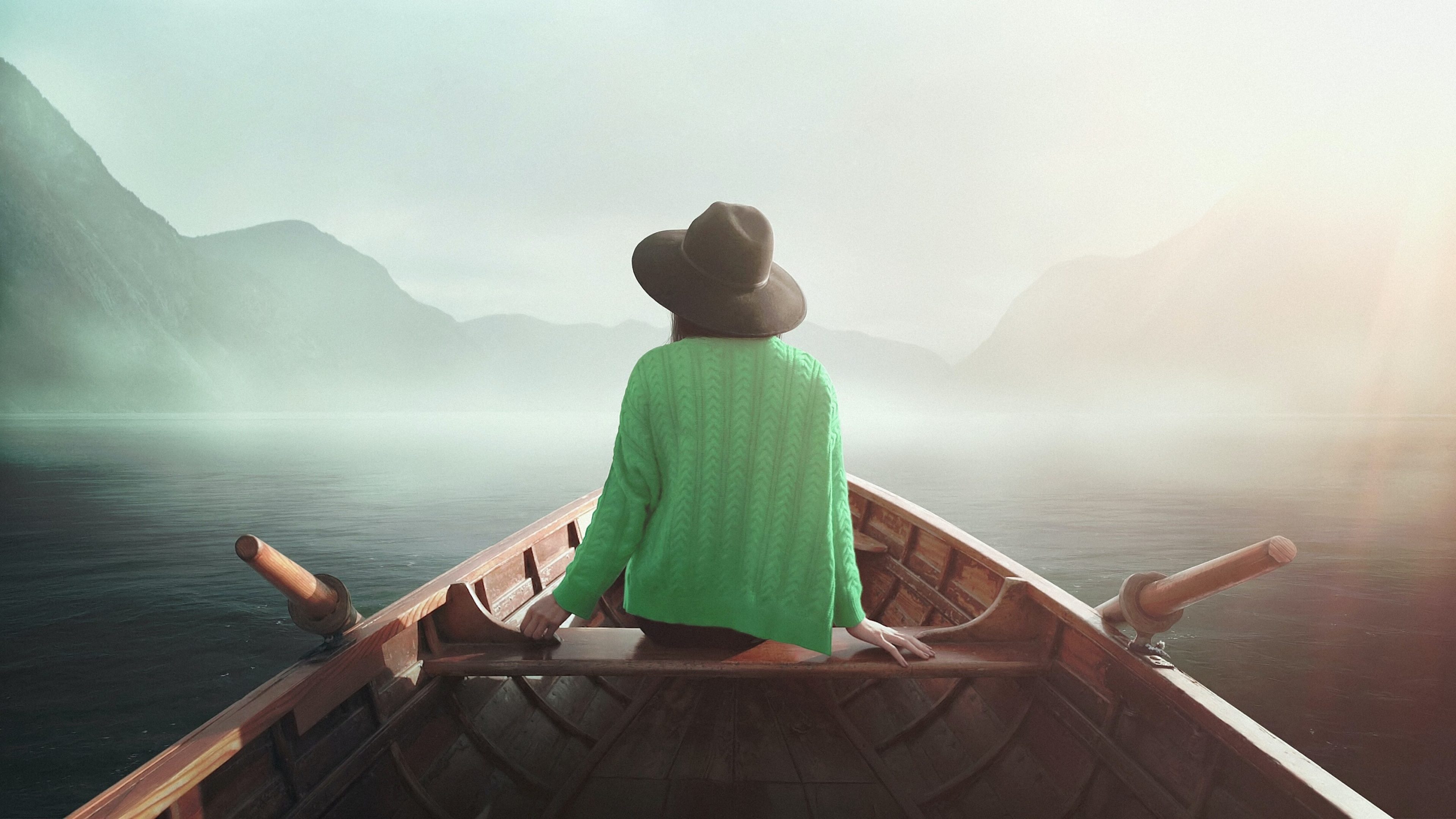 Eine Person mit einem schwarzen Hut und einem grünen Strickpullover sitzt in einem Holzboot und blickt auf einen nebligen See, umgeben von Bergen. Die Szene wirkt ruhig und mystisch.