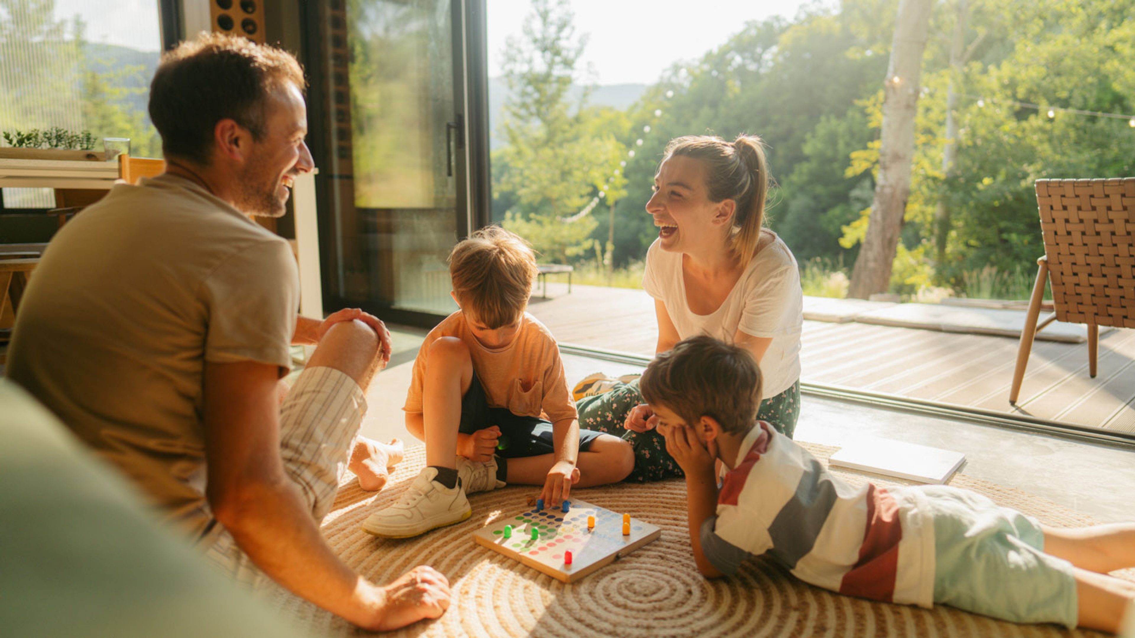 Zwei erwachsene Personen sitzen mit zwei Kindern am Boden neben einer offenen, lichtdurchfluteten Terrasse. Die Familie spielt «Mensch ärgere dich nicht» auf einem farbigen Brett. Die zwei Erwachsenen sehen sich lachend an und die zwei Kinder schauen fokussiert auf das Spielbrett.