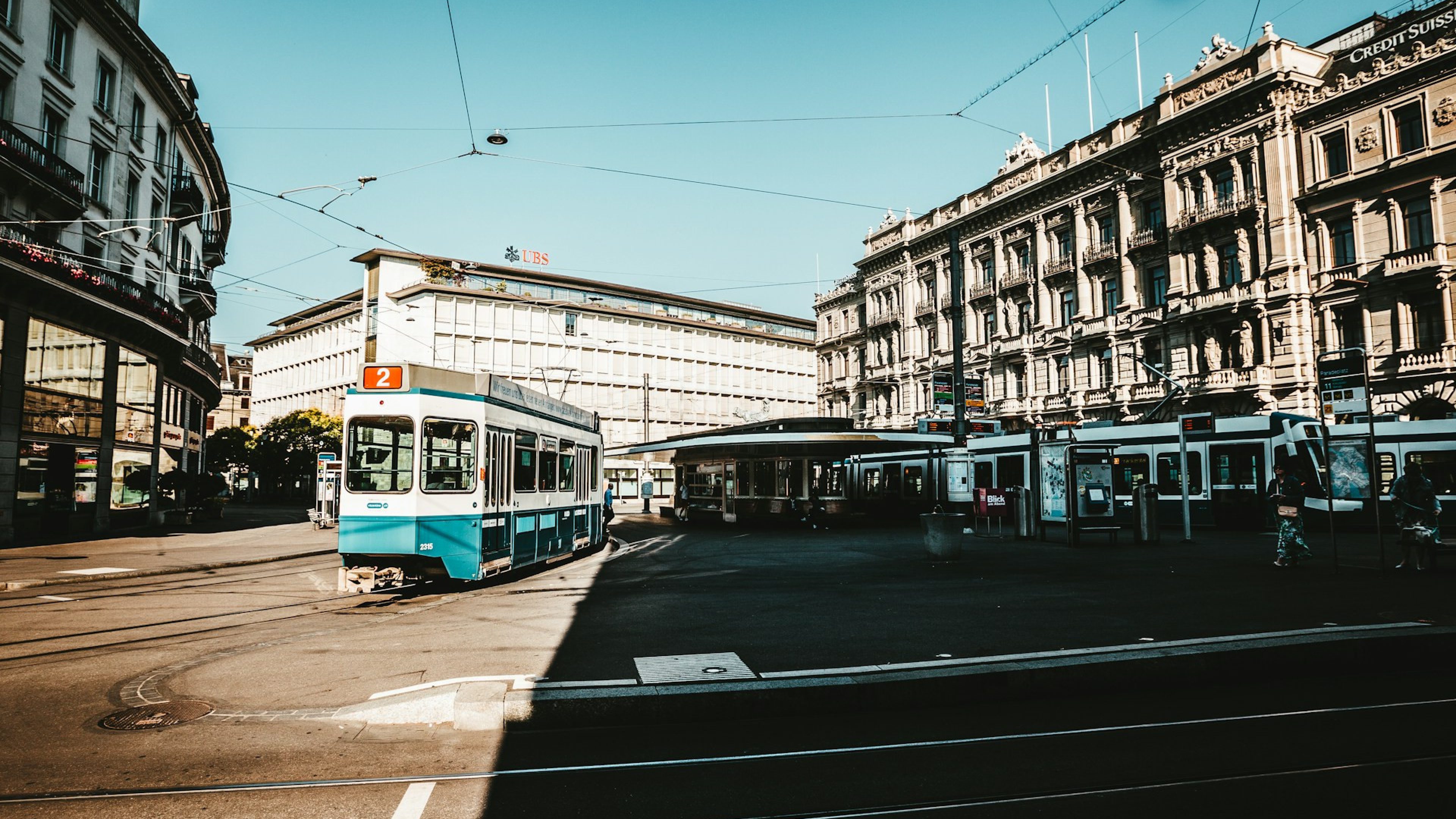 Strassenszene am Paradeplatz in Zürich mit einer vorbeifahrenden blauen Tram der Linie 2, umgeben von historischen Gebäuden und Bankenfassaden bei klarem, sonnigem Wetter. Das Bild zeigt die zentrale Verkehrsdrehscheibe mit Schienen, Haltestellen und ikonischer Architektur im Herzen der Zürcher Innenstadt.