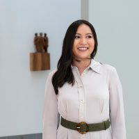 Eine Frau blickt mit einem strahlenden Lachen zur Seite. Sie trägt eine weisse Bluse  mit einem schwarzen Gürtel und hat mittellange schwarze Haare. Hinter ihr hängt eine kleine Holzskulptur mit drei laufenden Holzfiguren an einer weissen Wand.