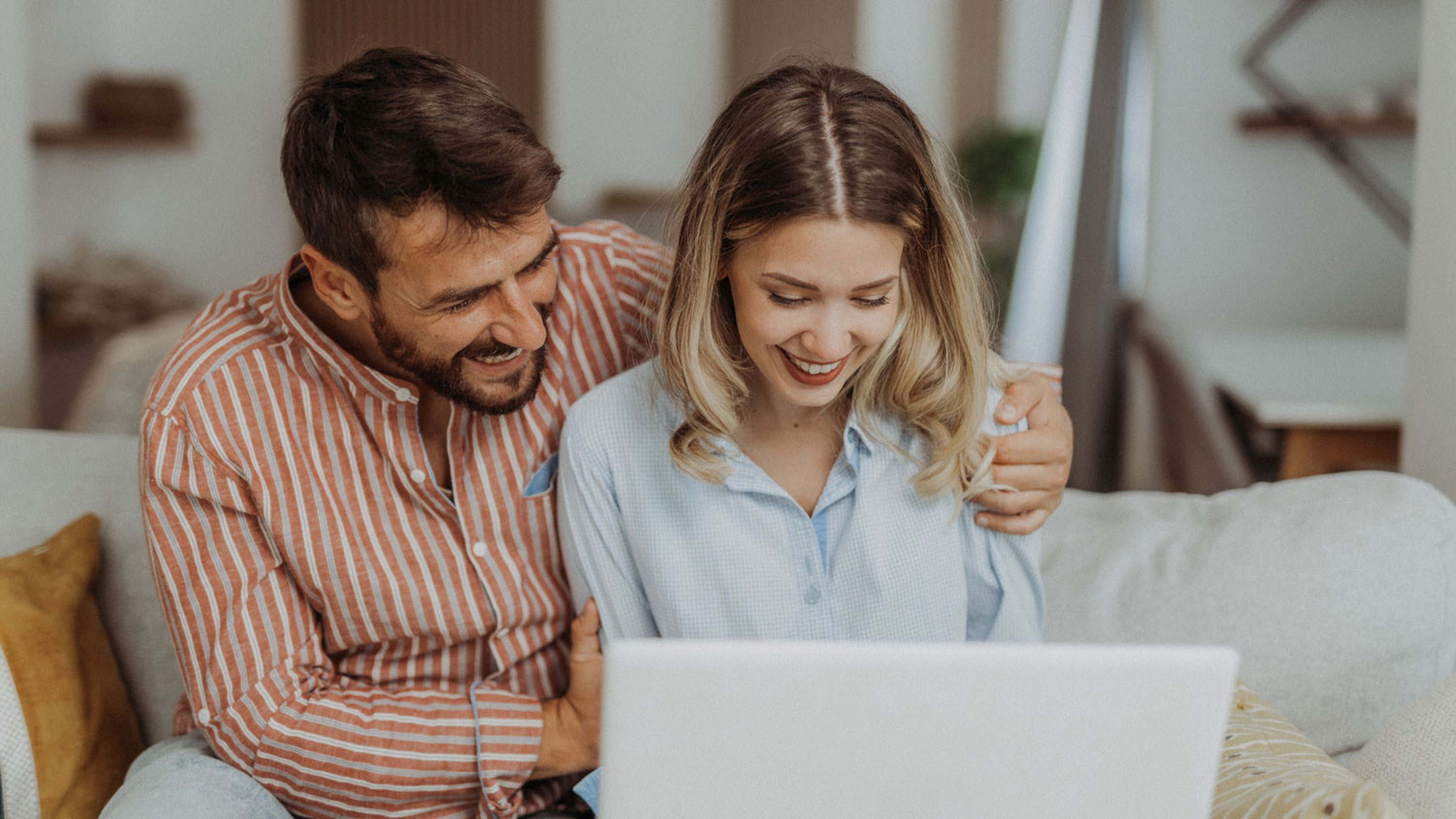 Eine Frau und ein Mann sitzen gemeinsam auf einem Sofa und schauen auf einen Laptop. Die Frau trägt eine hellblaue Bluse und hat blonde, schulterlange Haare. Der Mann trägt ein orange-weiss gestreiftes Leinenhemd, hat dunkle Haare und einen dunklen Bart. Im Hintergrund ist ein modernes Wohnzimmer mit Regalen und Möbeln zu sehen.