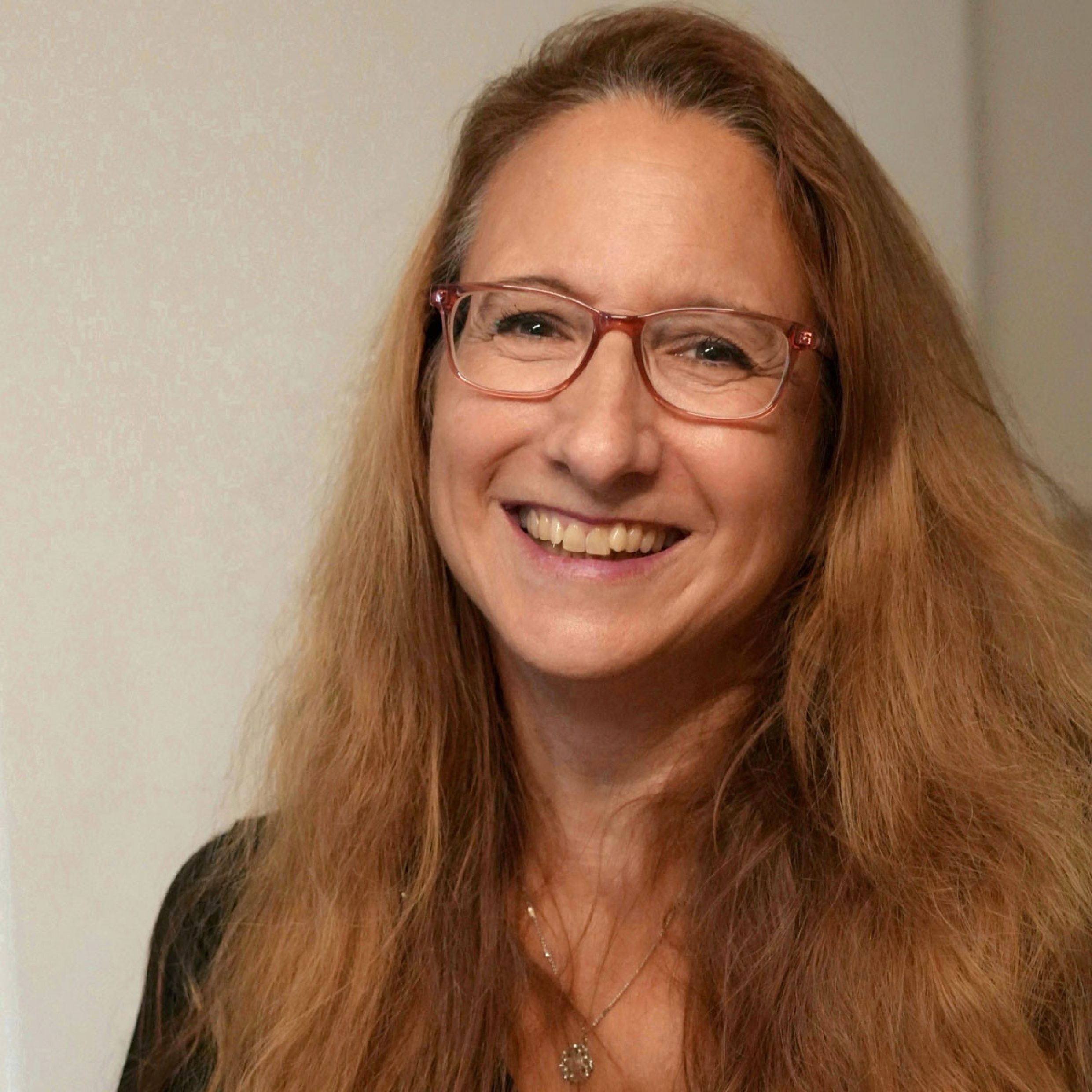 Ein Portrait einer Frau mit langem, rotblondem Haar und einer braunen Brille. Sie blickt in die Kamera und lächelt. Sie trägt ein schwarzes Oberteil und ist mittig im Bild platziert.