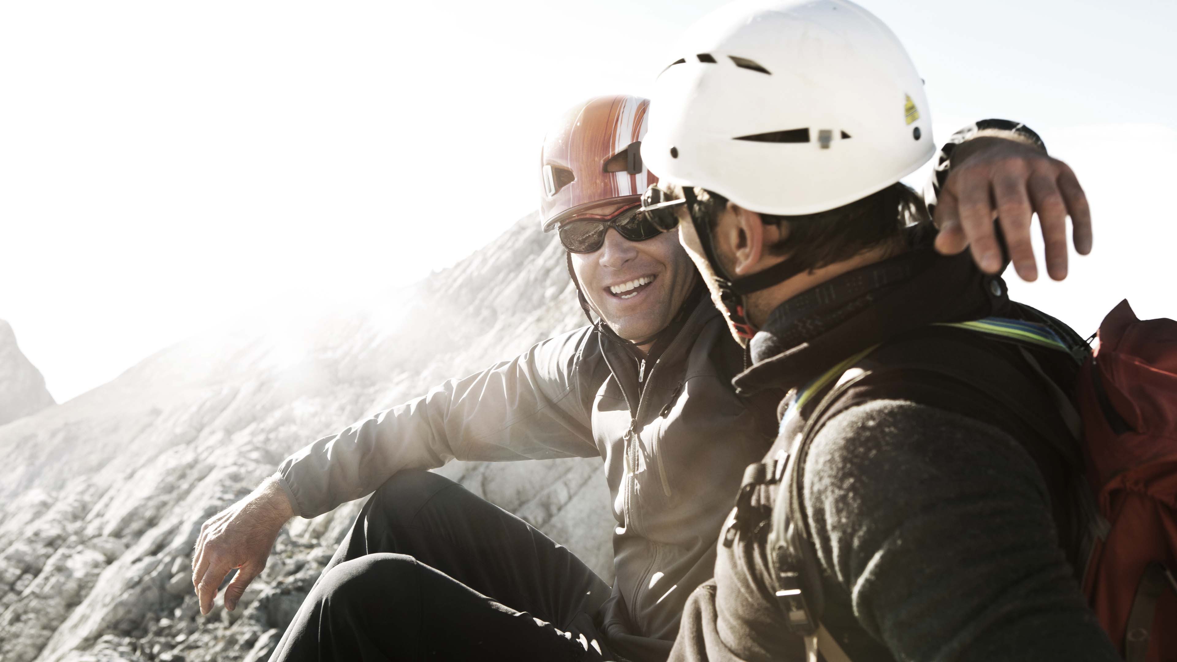 Zwei Personen sitzen auf felsigem Berggelände mit Helm und Ausrüstung, Sonne scheint, Gipfel im Hintergrund