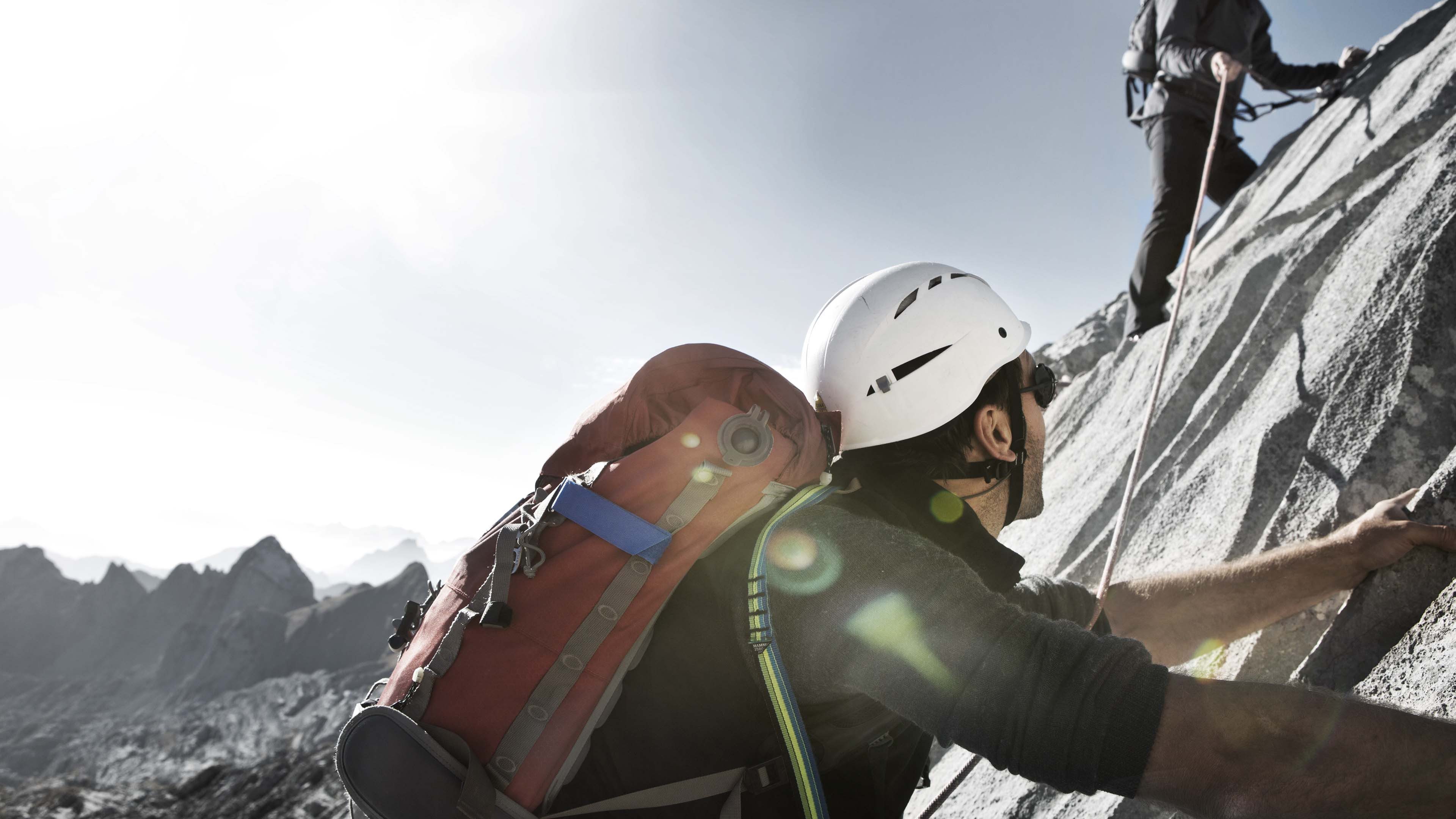 Zwei Kletterer steigen steile Felswand hinauf, mit Helm und Seilen gesichert, Sonnenlicht und Berggipfel im Hintergrund