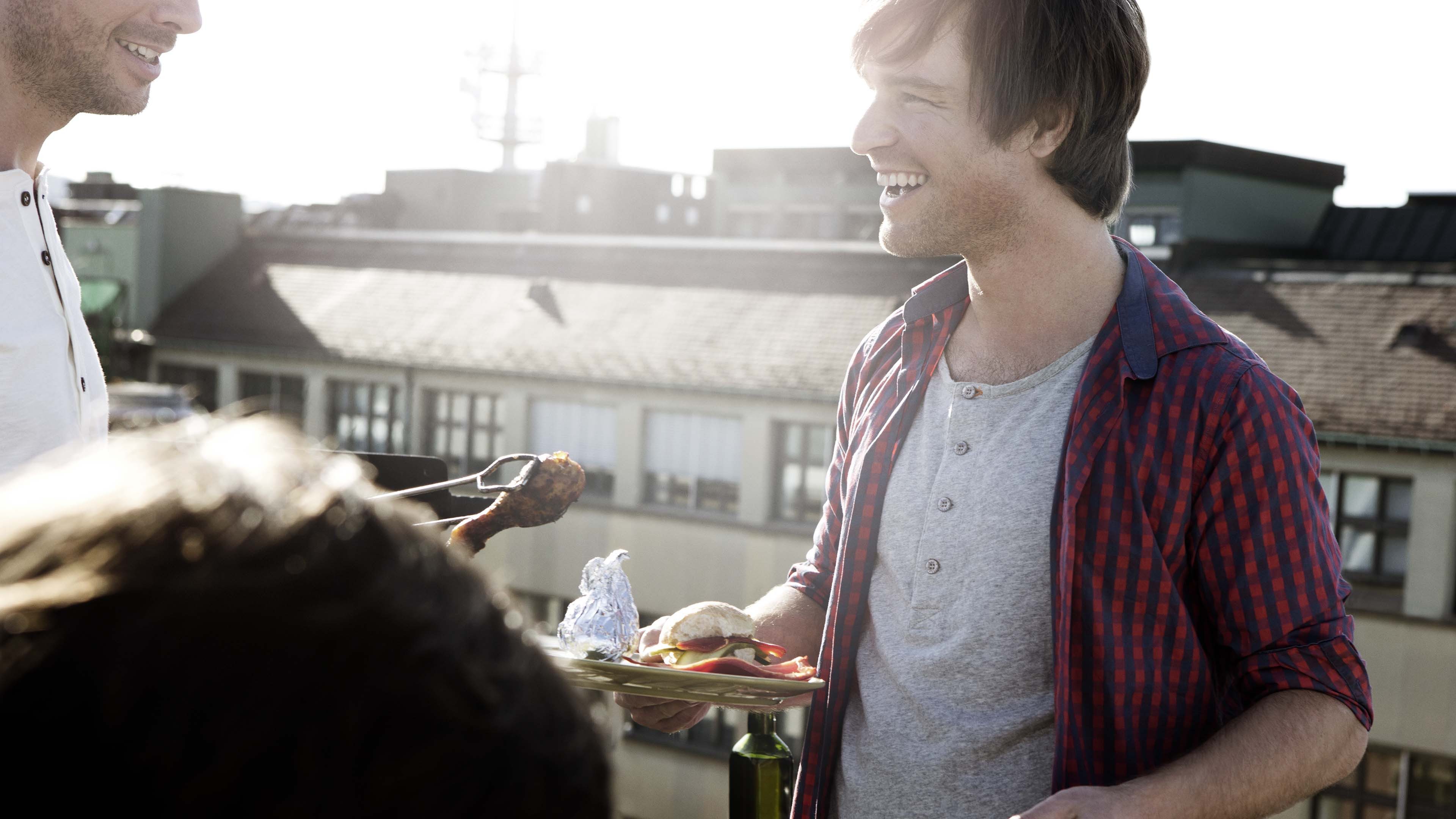 Gruppe auf Dachterrasse, eine Person mit Teller, eine mit Spieß, Gebäude und Himmel im Hintergrund