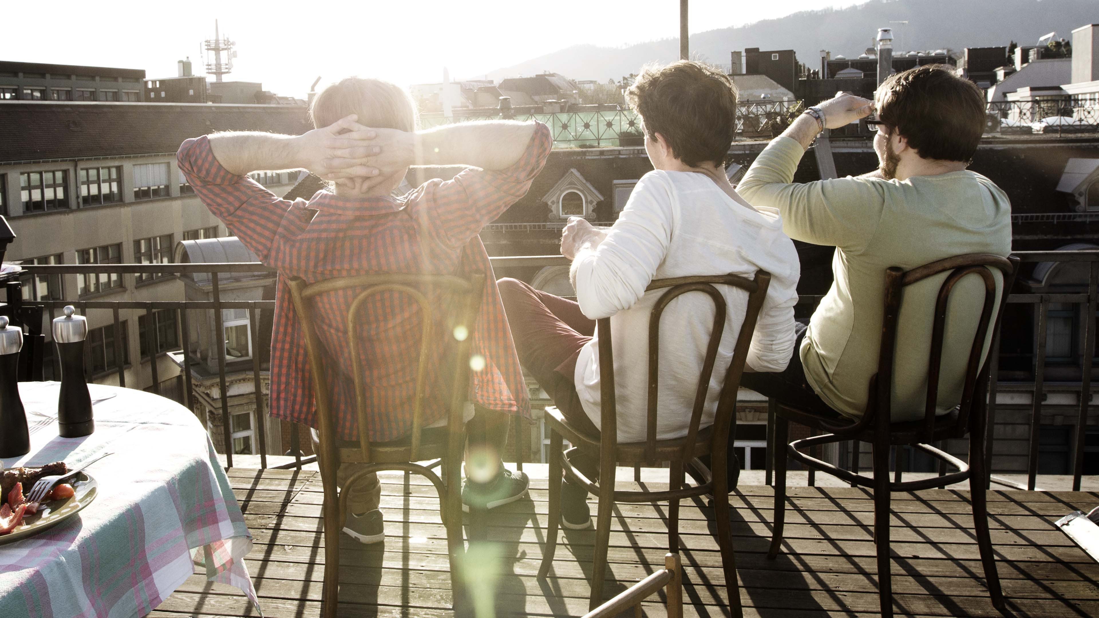 Drei Personen sitzen auf Dachterrasse und blicken in den Sonnenuntergang über Stadt und Hügel