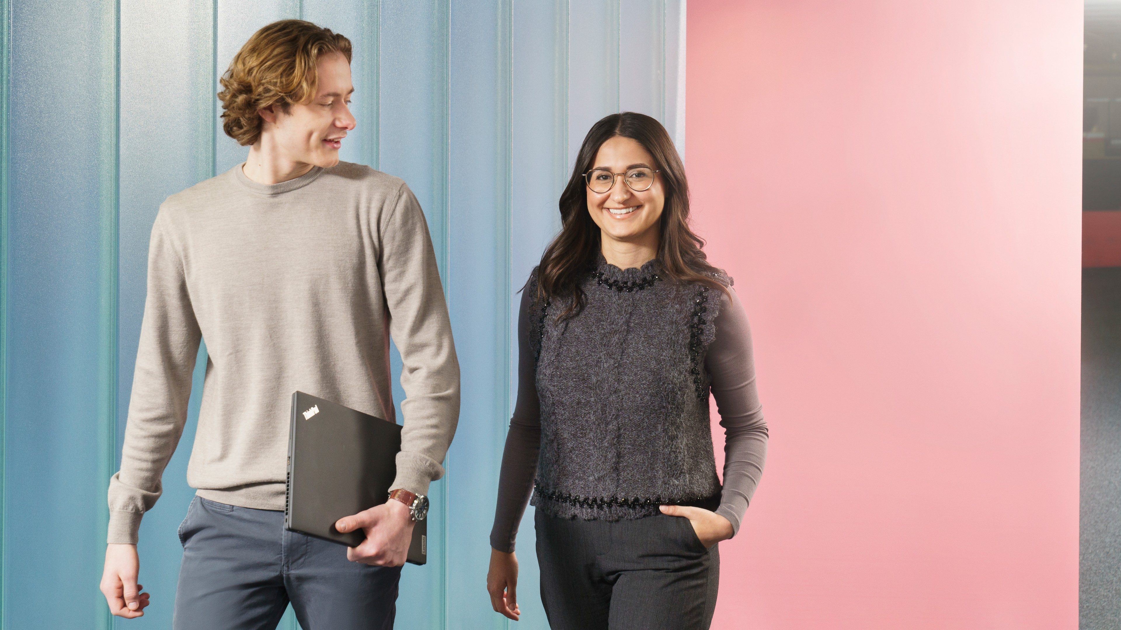 Zwei Future Minds gehen Seite an Seite. Die Frau steht rechts und lächelt direkt in die Kamera. Der Mann geht links von ihr, hält einen Laptop zusammengeklappt in der Hand und spricht seitlich abgewandt mit der Frau.