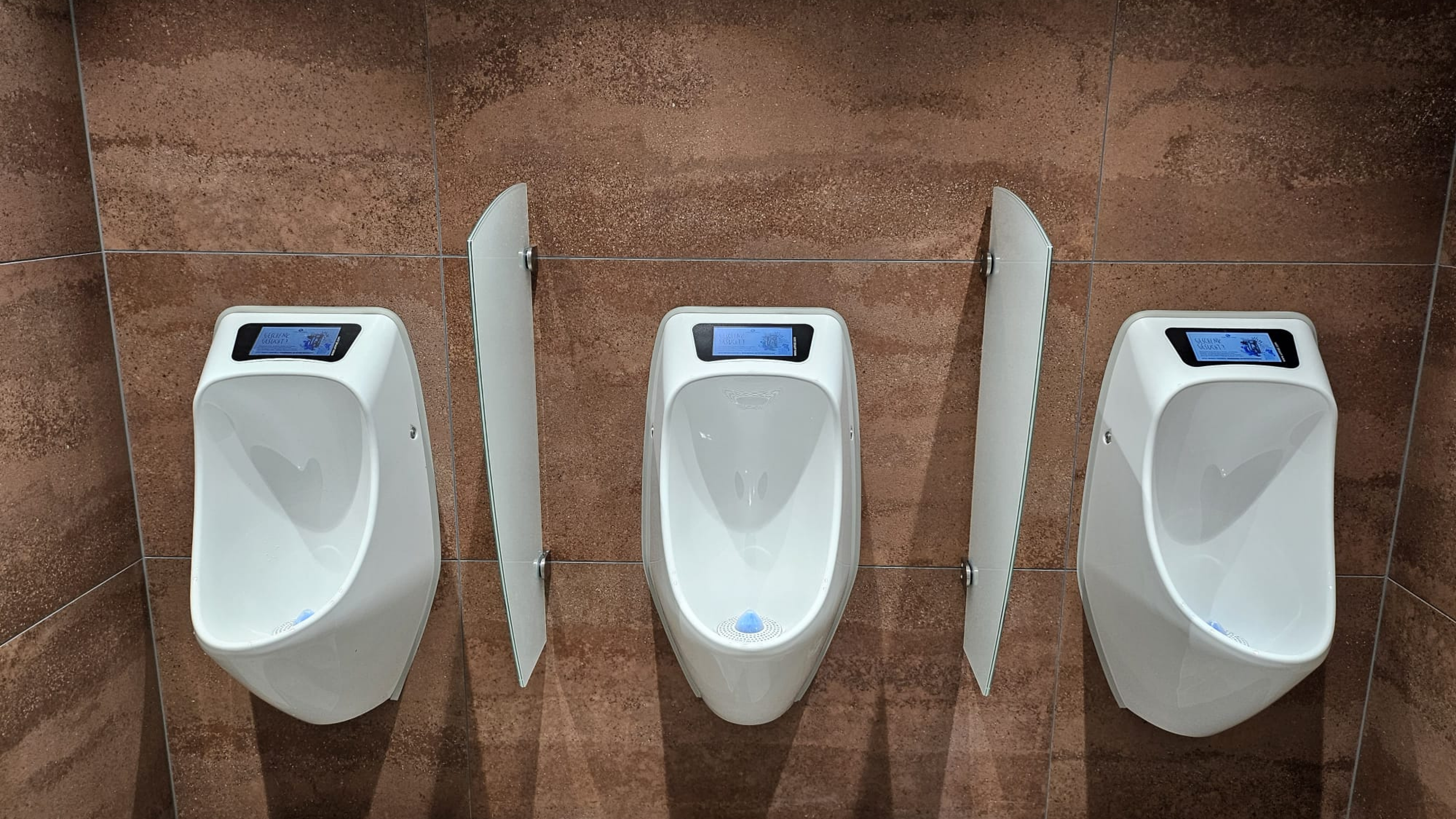 Drei weisse Urinale mit Display, montiert an einer braunen Fliesenwand und durch Glaswände getrennt – modern und sauber.