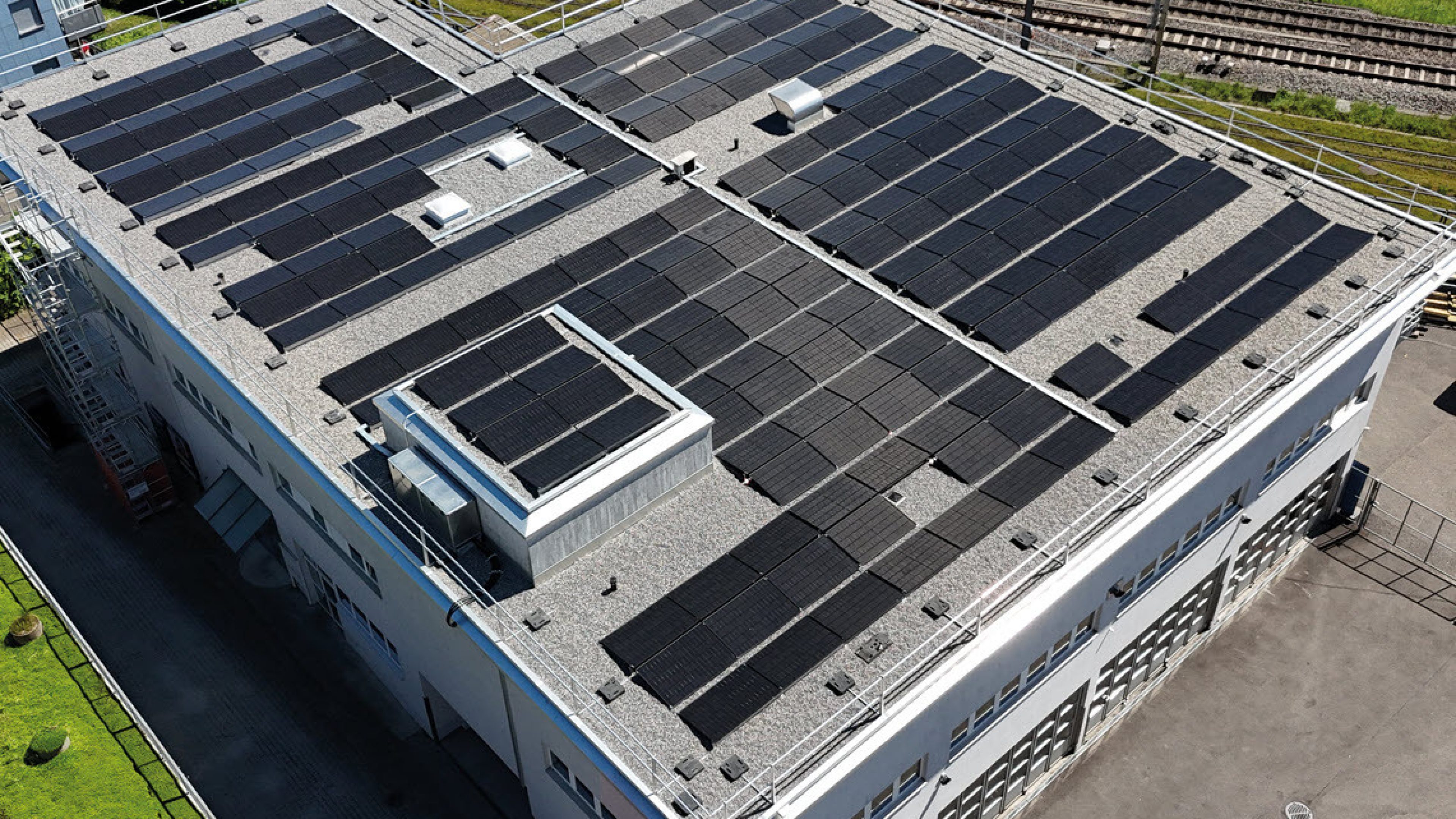 Luftaufnahme des grossen Firmengebäudes mit Solarpanels auf dem Dach, umgeben von Gleisen, Grünflächen und parkierten Fahrzeugen bei klarem Himmel.