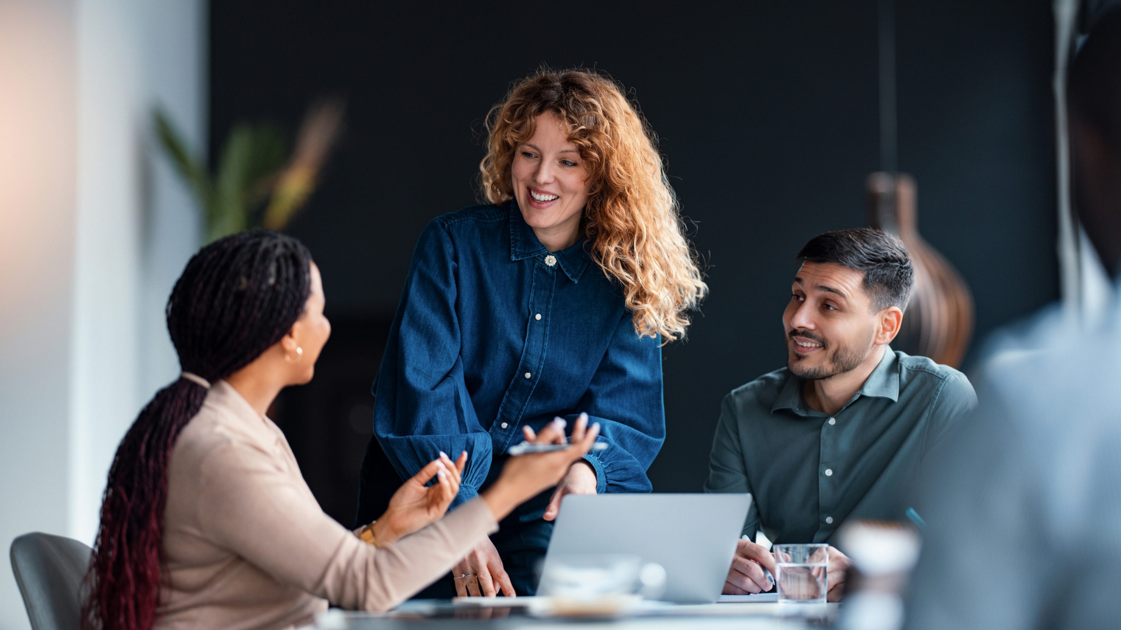 Drei Personen sind um einen Tisch in einem modernen Büro versammelt. Zwei Personen sitzen, die in der Mitte steht. Sie diskutieren gemeinsam über ein Projekt. Auf dem Tisch liegt ein Laptop, daneben steht ein Glas Wasser. Im Hintergrund ist eine Pflanze sichtbar.