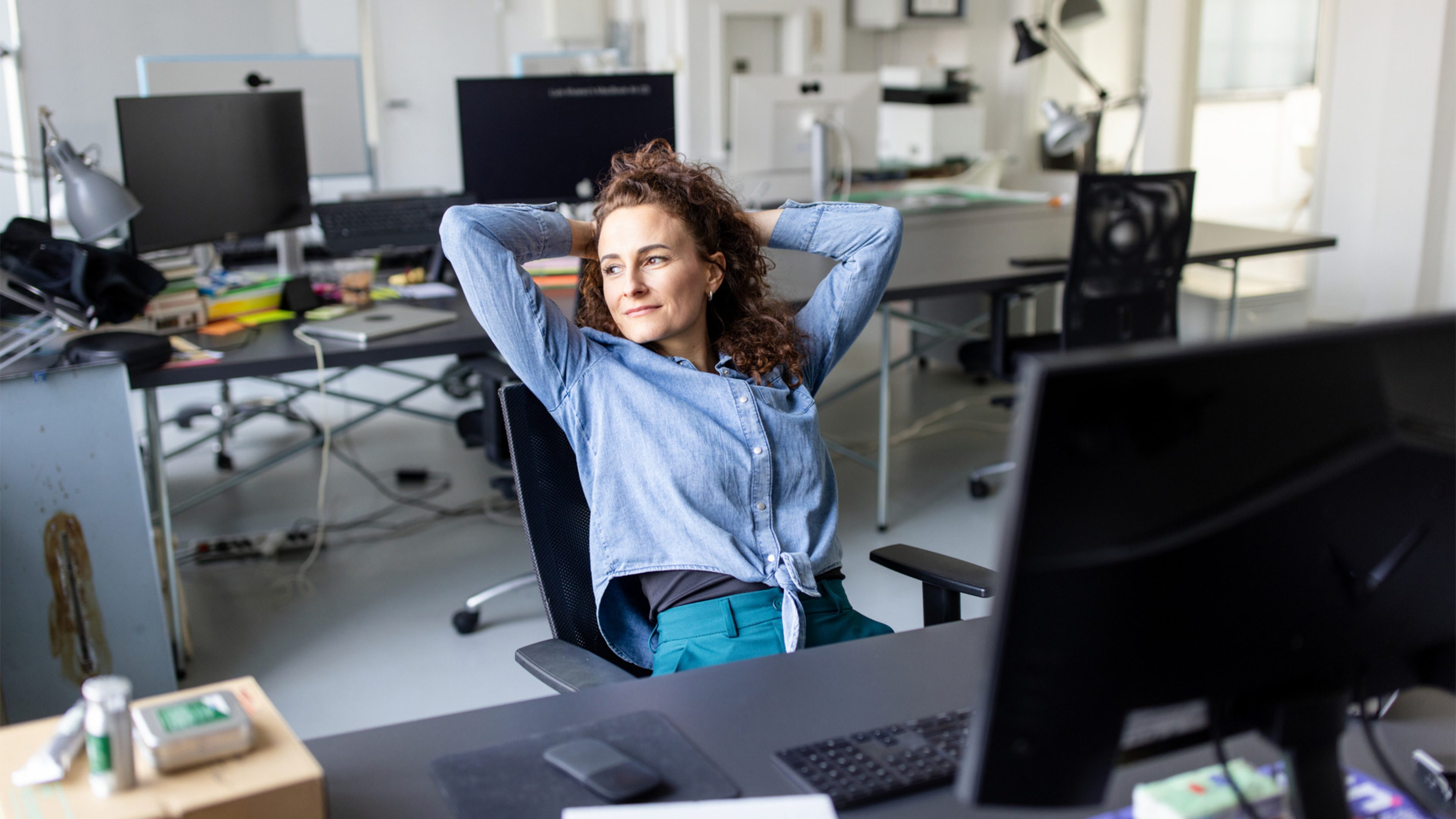 Eine Frau mit blauer Jeansbluse und dunklen Locken sitzt in einem Bürostuhl. Sie lehnt sich nach hinten mit ihren Armen über dem Kopf. Ihr Blick ist nach links gerichtet und sie ist mittig im Bild positioniert. Vor ihr ist ein Bürotisch mit Tastatur, Maus und Monitor leicht verschwommen sichtbar. Hinter ihr ist eine Büroumgebung zu erkennen. Der Raum ist ausgestattet mit weiteren Bürotischen, schwarzen Monitoren, Tischlampen und Bürostühlen. Die Wände des Raumes sind weiss.  