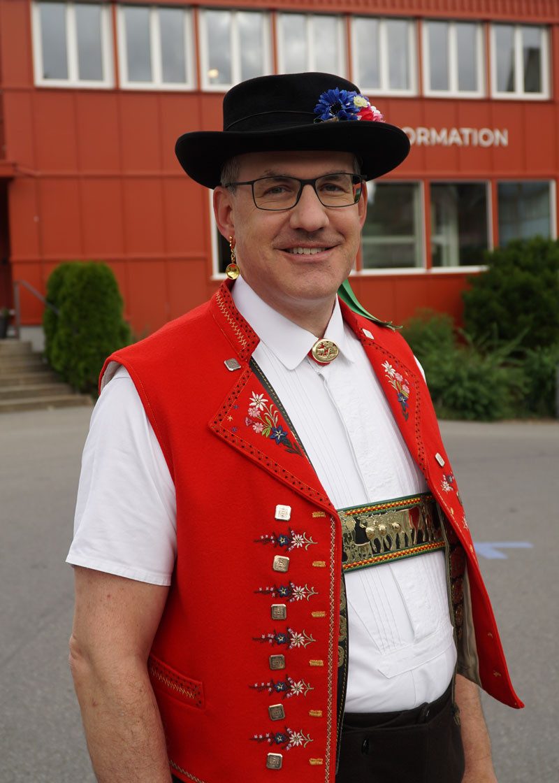 Ein Mann in traditioneller Appenzeller Kleidung mit roter, verzierter Weste, weissem Hemd und schwarzem Hut steht vor einem roten Gebäude mit der Aufschrift «Tourist Info».