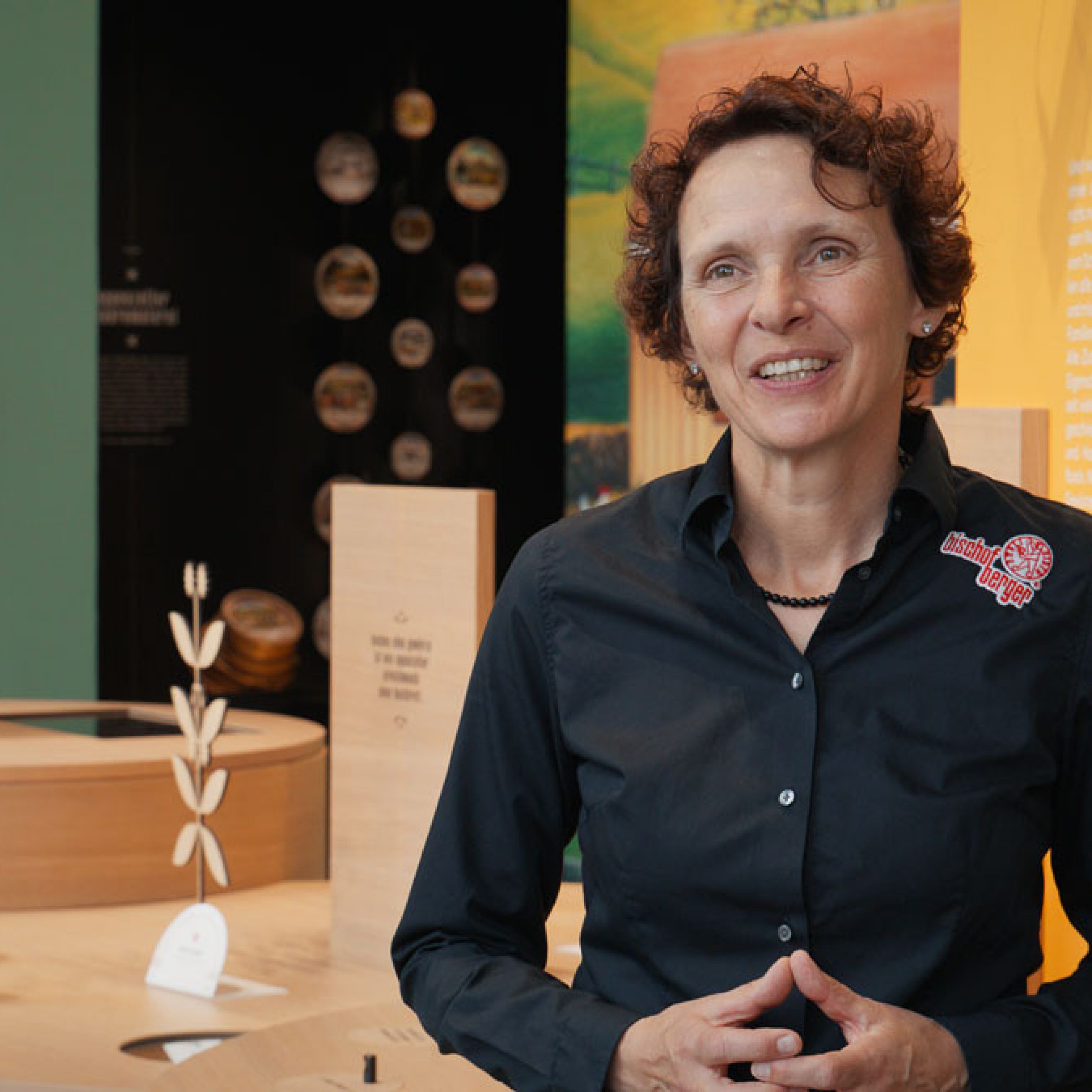 Eine Frau steht in einem Ausstellungsraum mit Holzständen, Gläsern und Infotafeln. Im Hintergrund ist ein farbiges Wandbild mit Gebäuden und Naturmotiven zu sehen. Die Frau trägt eine schwarze Bluse mit dem Logo der Bischofberger AG.