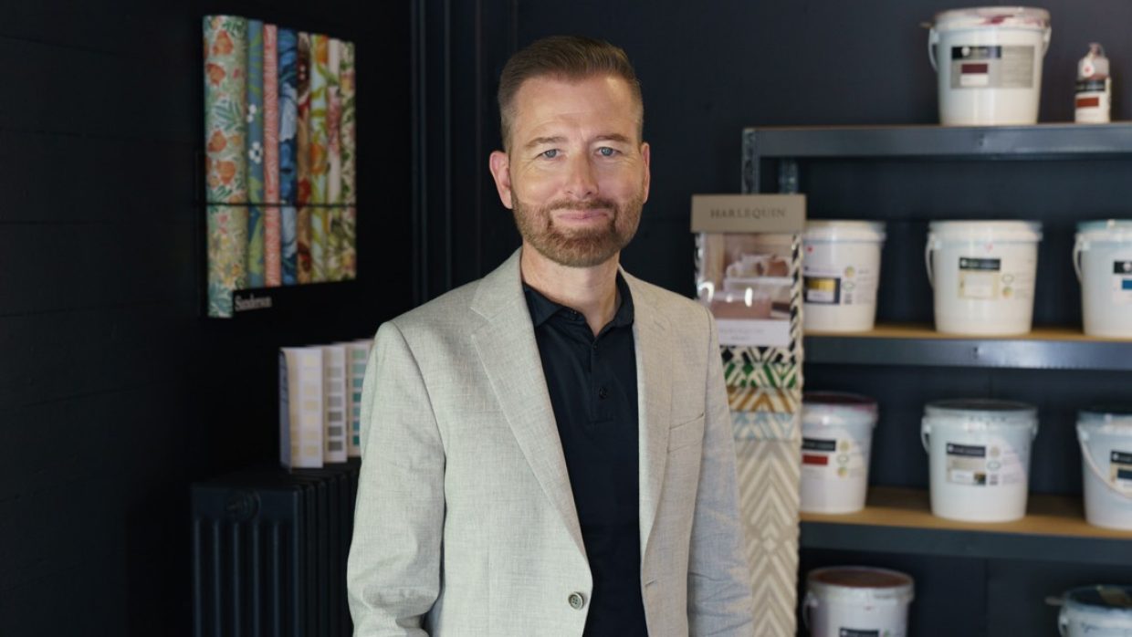 Hinter einem Mann mit grauem Blazer zieren Farb- und Tapetenmuster die Wände eines Showrooms. Die Muster dienen sowohl als Ansichtsexemplare als auch als Dekor.