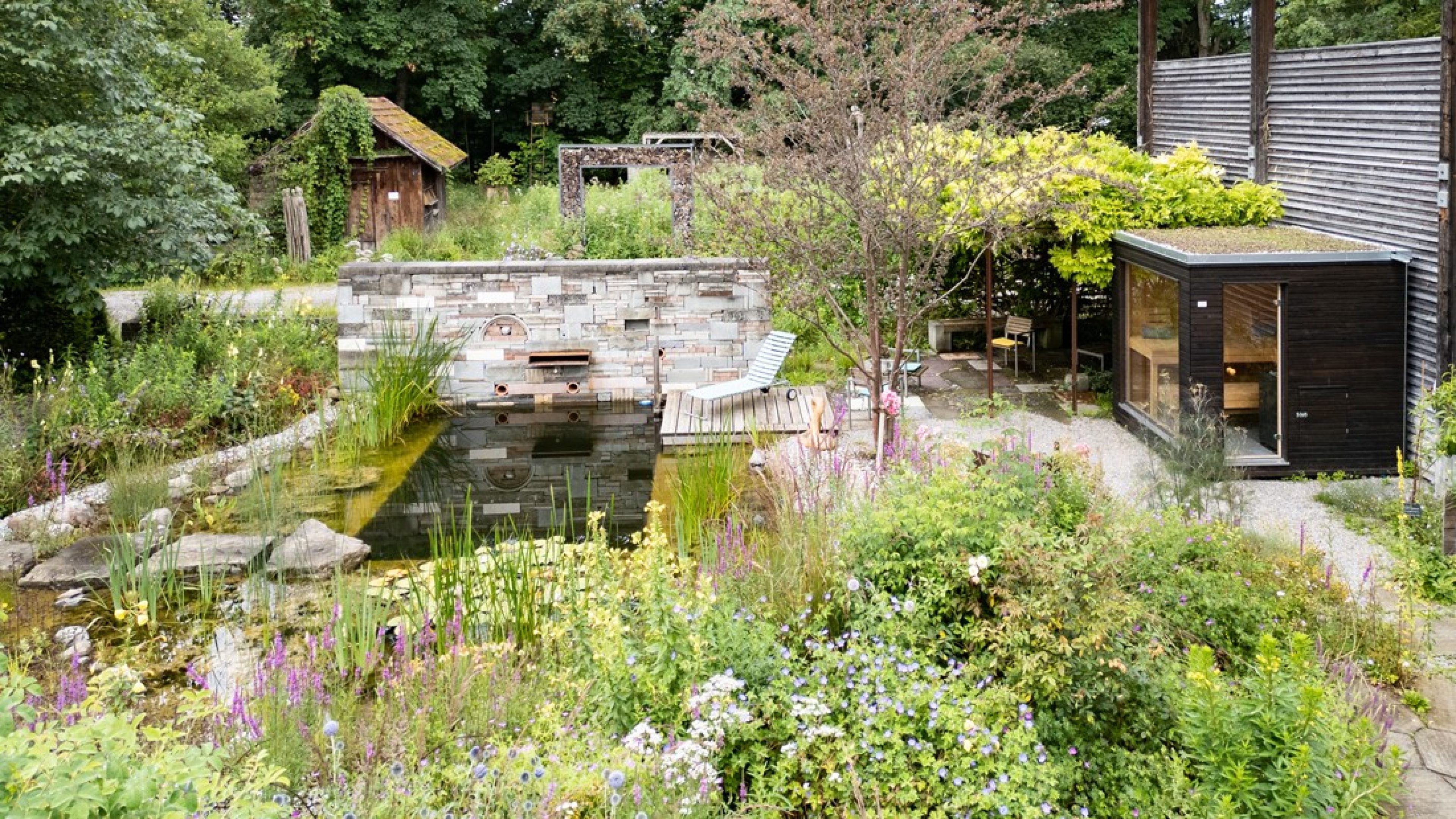 Ein Einblick in einen Garten voller verschiedener Grünpflanzen, die mit blauen und violetten Blumen durchsetzt sind. Mitten im Garten ist ein kleiner Teich mit einem Sitzplatz, der mit einem Liegestuhl zum Entspannen einlädt.