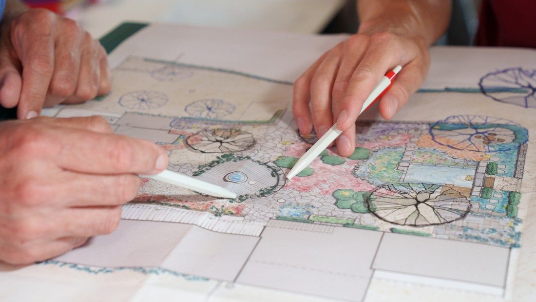 Zwei Personen besprechen etwas anhand des Gartenplans. Man blickt auf ihre Hände, die mit einem Kugelschreiber auf einen bepflanzten Kreisel im Garten zeigen.