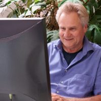 Ein Mann in einem blauen Hemd blickt auf den Bildschirm eines Computers, während er etwas tippt. Hinter ihm ist eine Monstera-Pflanze mit grossen, dunkelgrünen Blättern. 