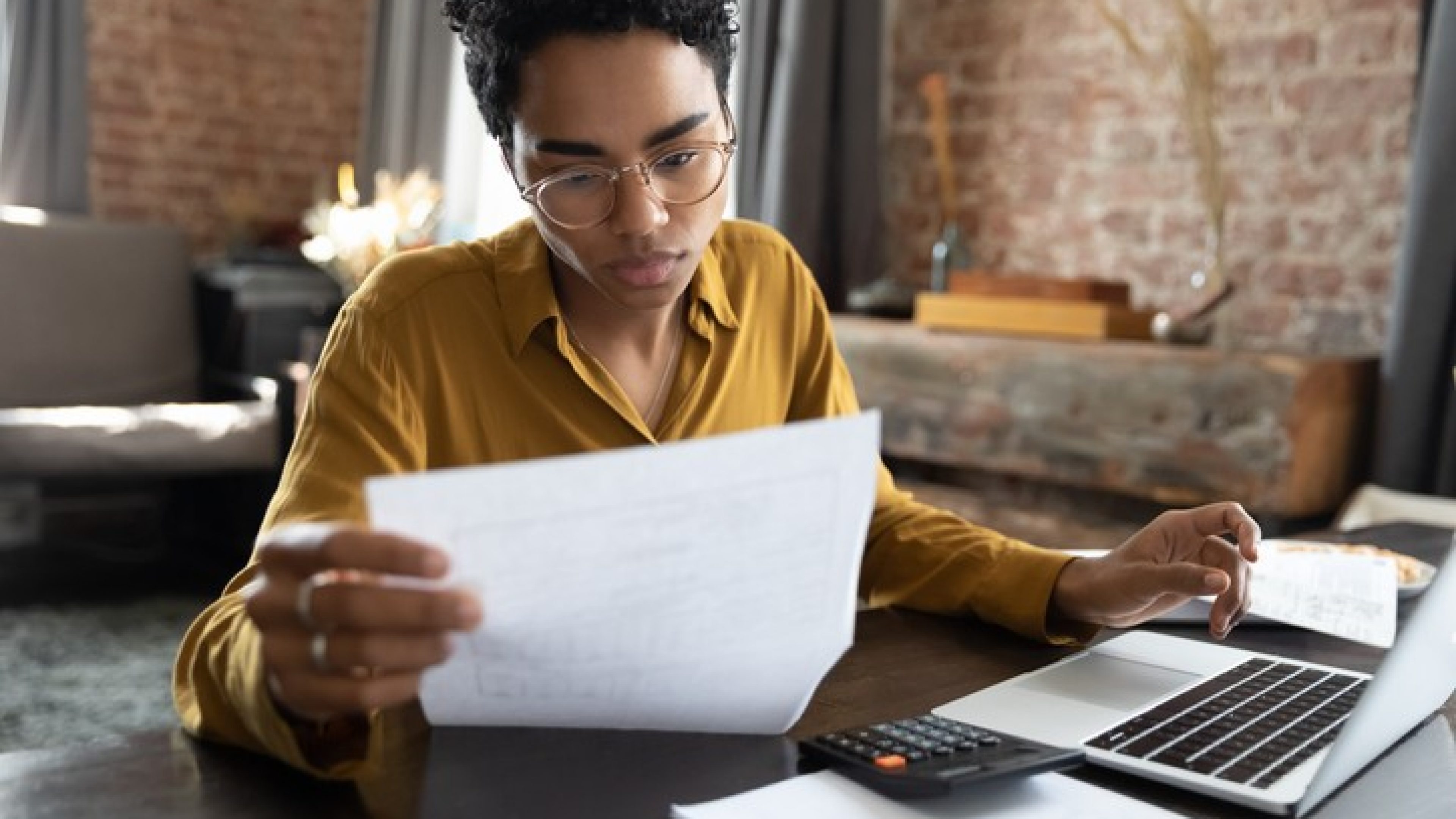 Focused young African American woman in eyeglasses looking through paper documents, managing business affairs, summarizing taxes, planning future investments, accounting alone at home office.