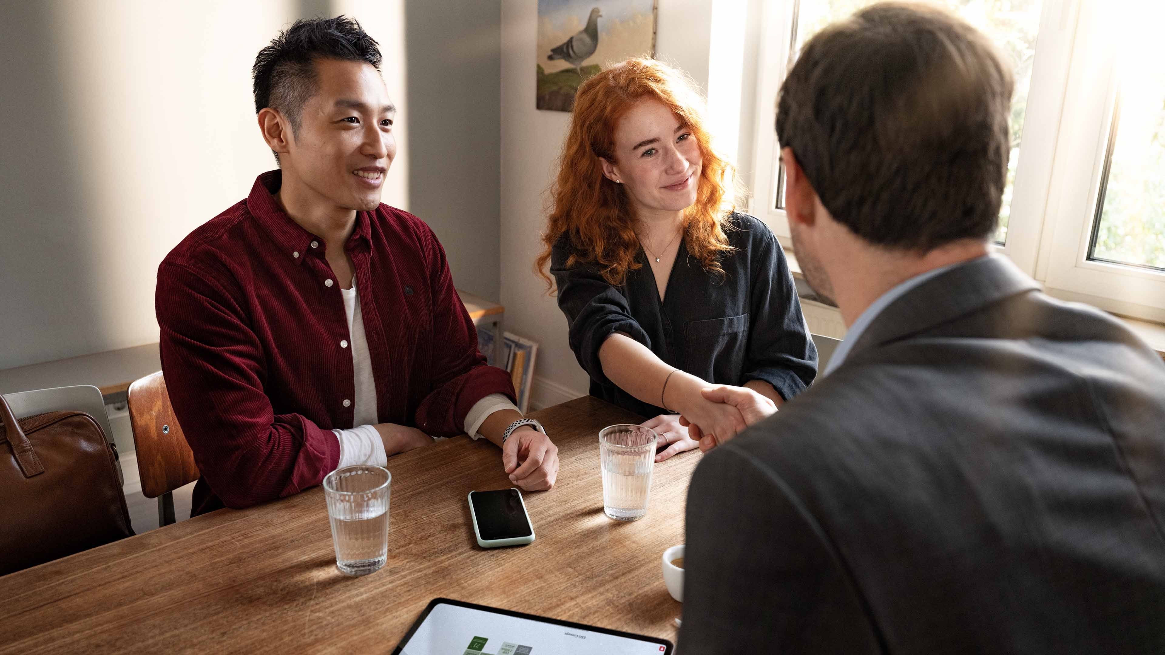 An insurance counsellor is advising a couple (a man and a woman) in a meeting room. He and the woman are greeting each other and shaking hands.