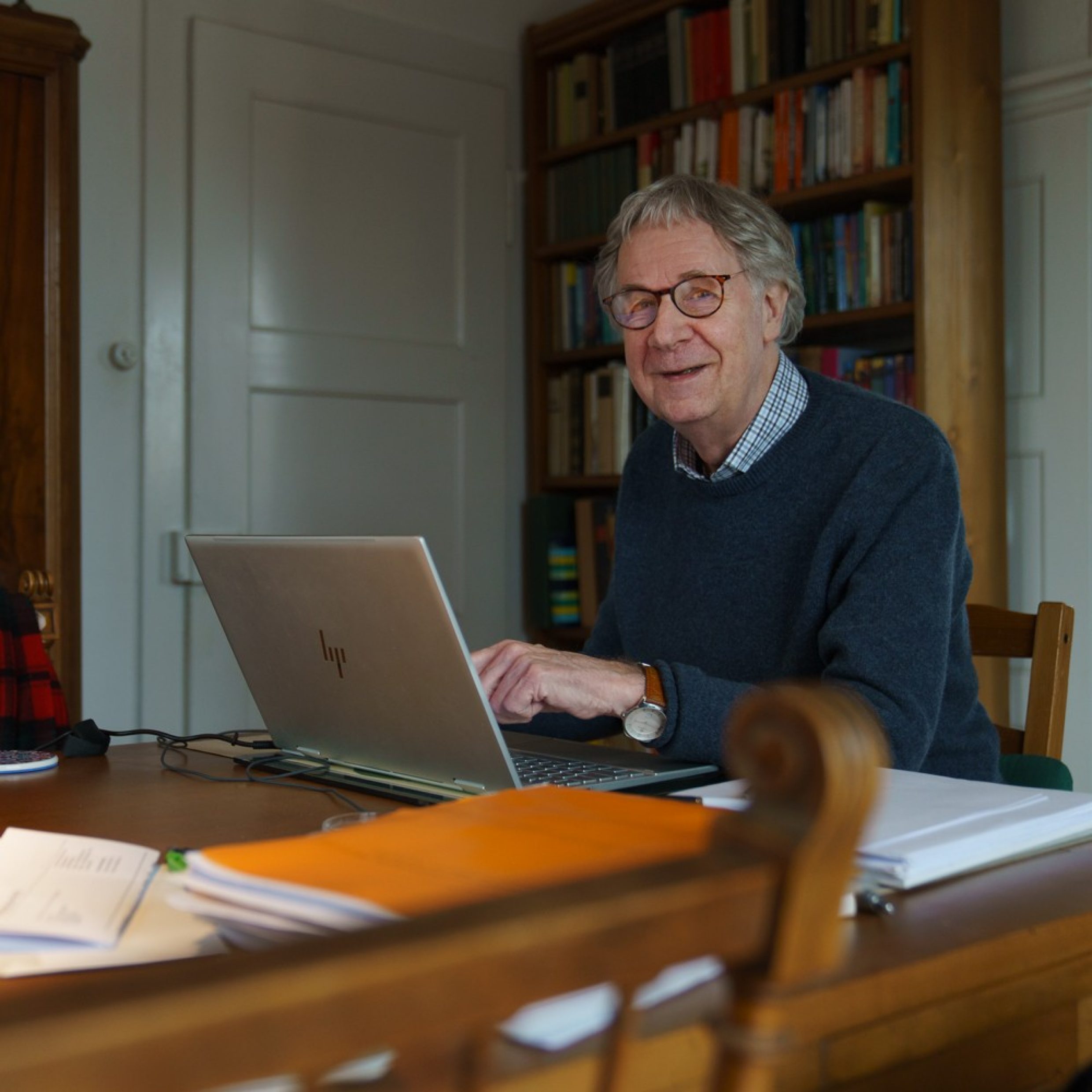 An older man sits at a table in front of a laptop, pointing to something on the screen. He smiles at the camera. The man has grey hair and is wearing glasses and a blue sweater.