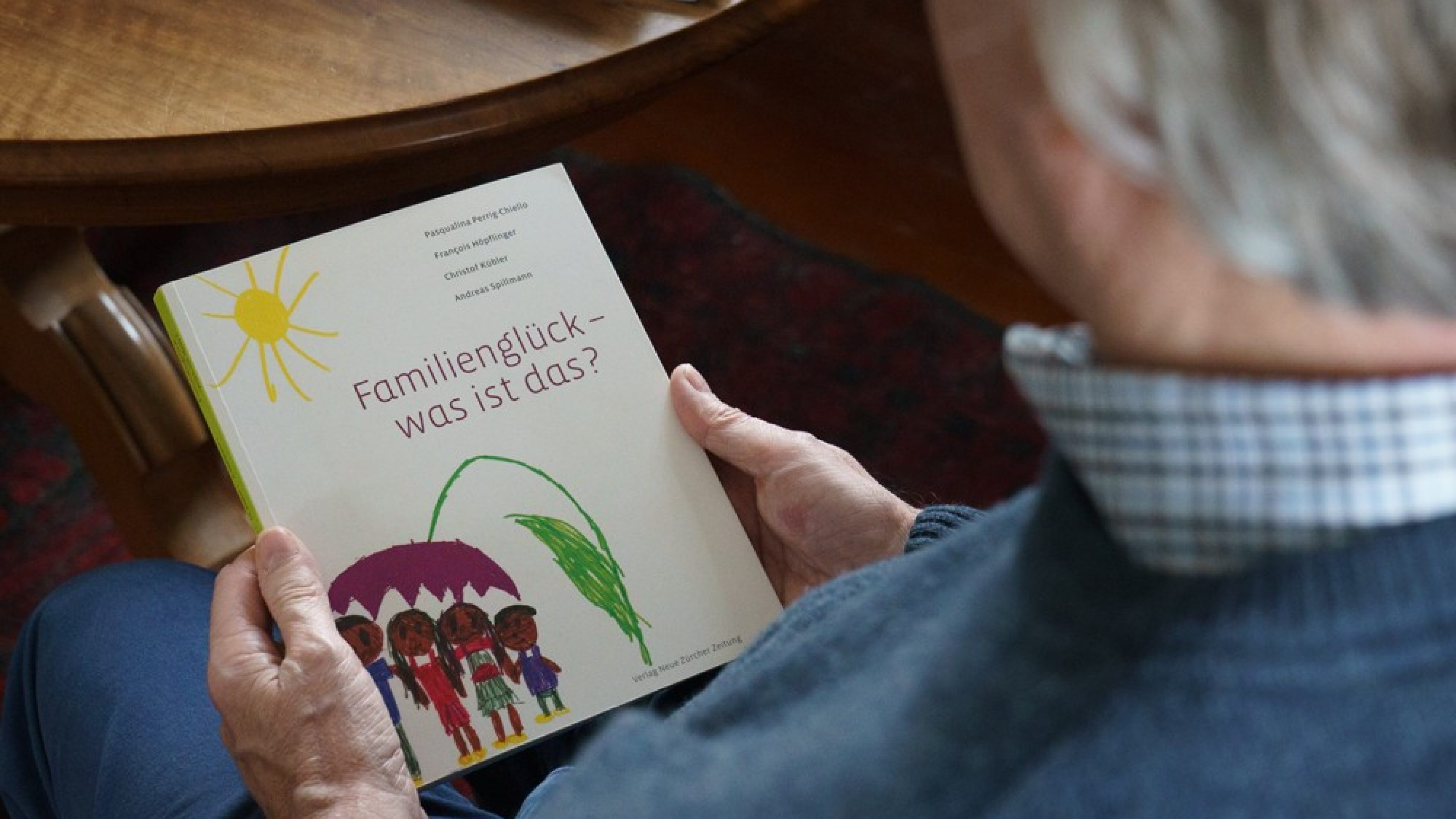 Blick über die Schulter eines älteren Mannes. Dieser hält ein Buch in der Hand mit dem Titel «Familienglück – was ist das?».