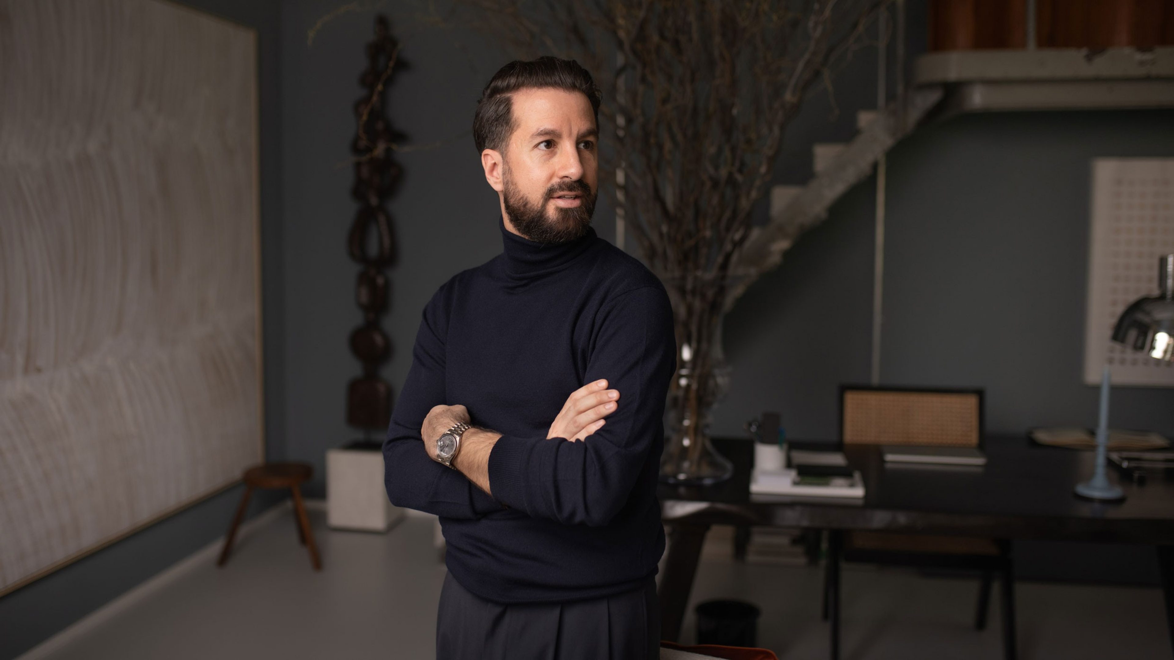 A man in a dark polo-neck pullover stands with his arms folded in a stylishly furnished office. He wears a watch and looks thoughtfully to the side. A large desk with minimalist office accessories is visible in the background. A staircase leads upstairs, next to which an abstract work of art and a large, dry branch in a vase serve as decorations. The atmosphere is modern and professional.