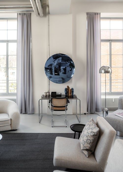 A bright room with high ceilings and large windows. In the centre, a round, blue mirror hangs on the wall, which sets a striking accent. There is a desk and a chair in front of the mirror. Sofas are arranged around it.