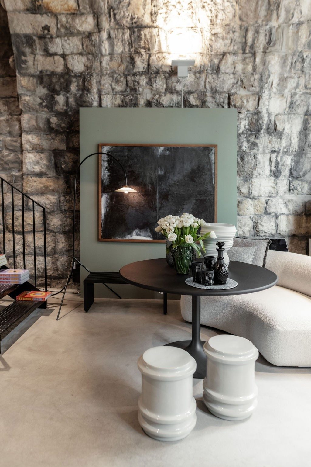 On a stone wall stands a loden green decorative wall with a square abstract picture. In front of it, on a bright carpet, stands a round black table with decorative items and white flowers in a vase. Around this table is a white couch and two white stools reminiscent of chess pieces.