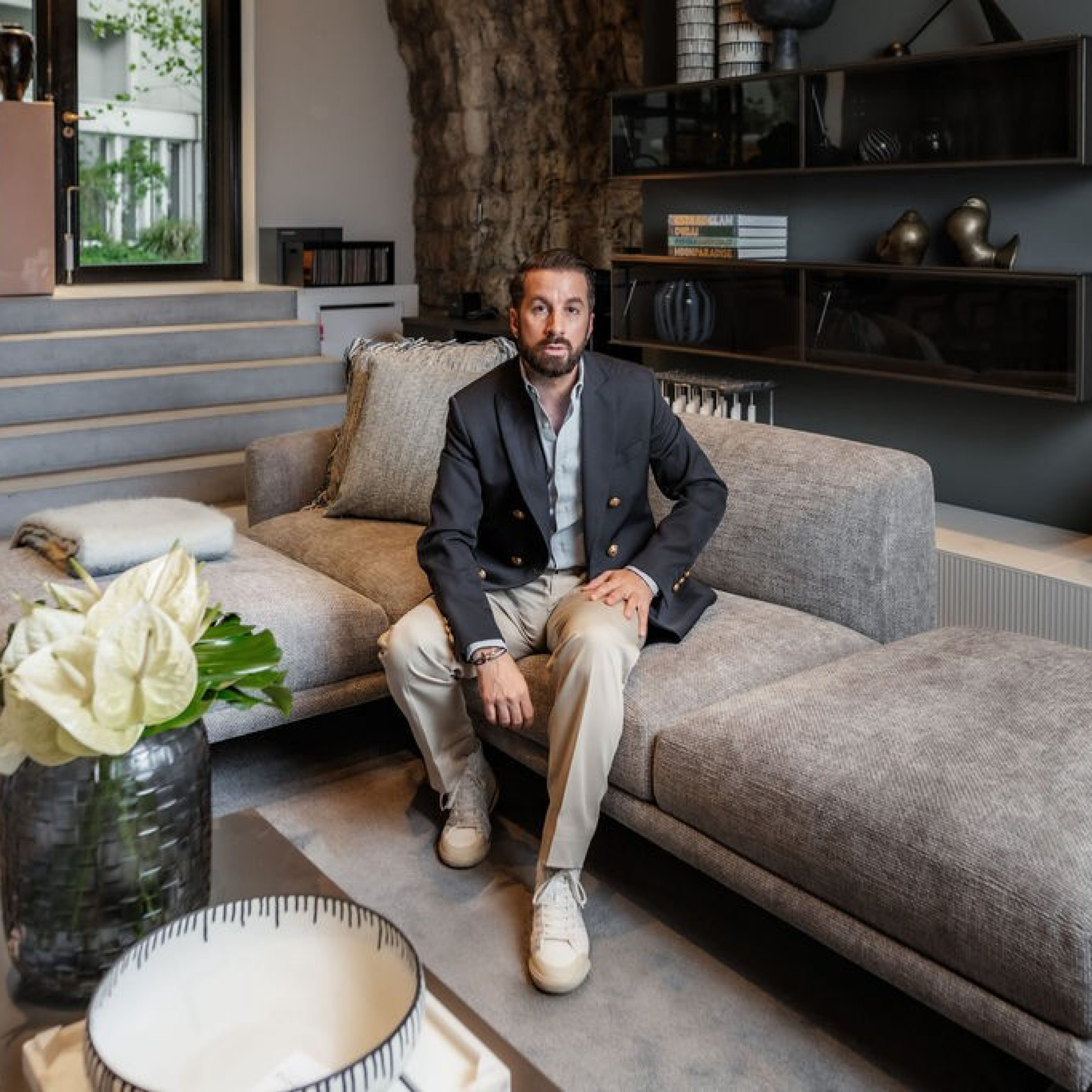 A dark-haired man, dressed in a denim shirt and with folded arms, looks into the camera. He is standing in a living area, with sofas and a TV behind him.