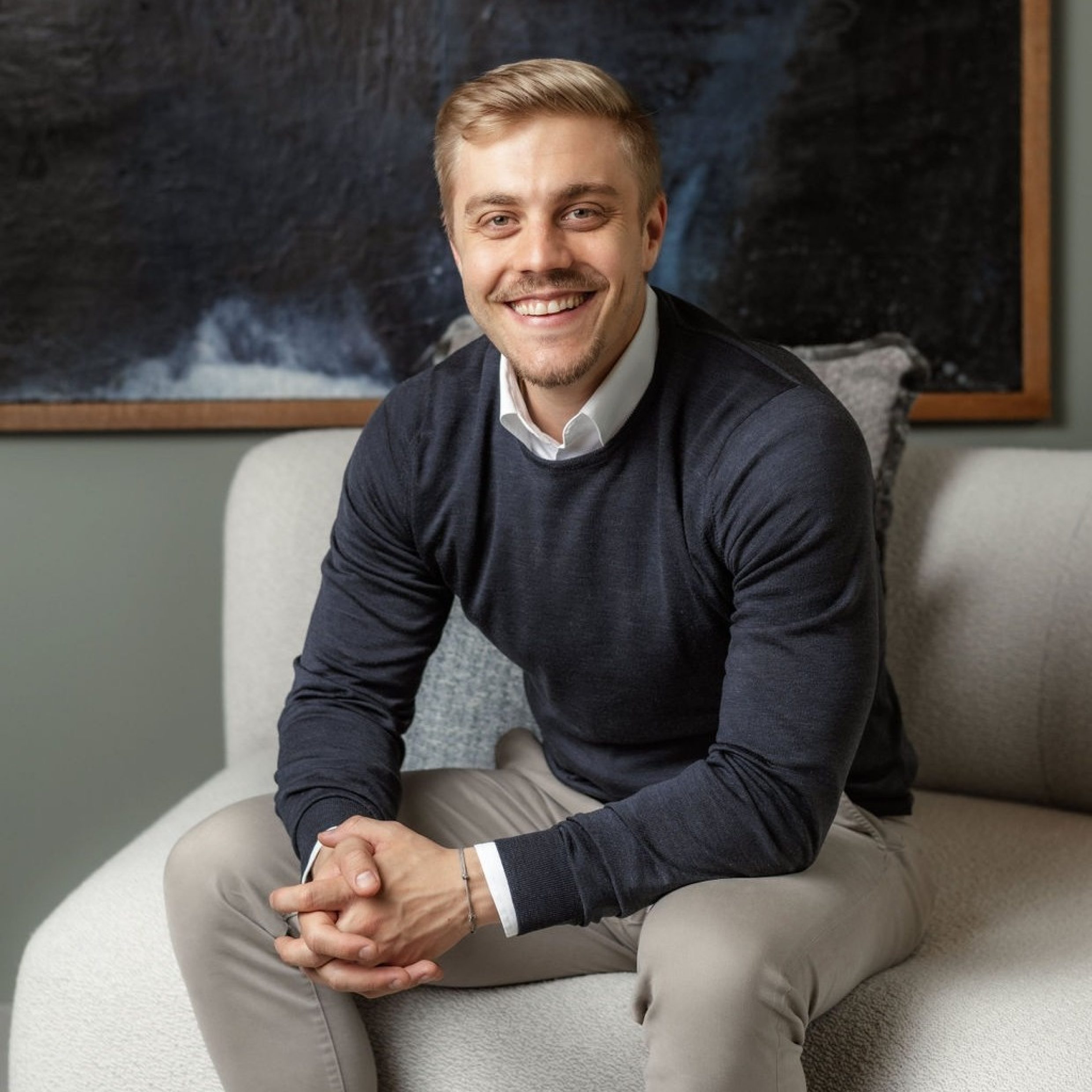 Man smiling on a couch with a sunny appearance. His dark blue sweater matches the abstract image in the background.