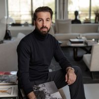 A man in a dark outfit looks into the camera. An elegantly furnished living area is visible behind him.
