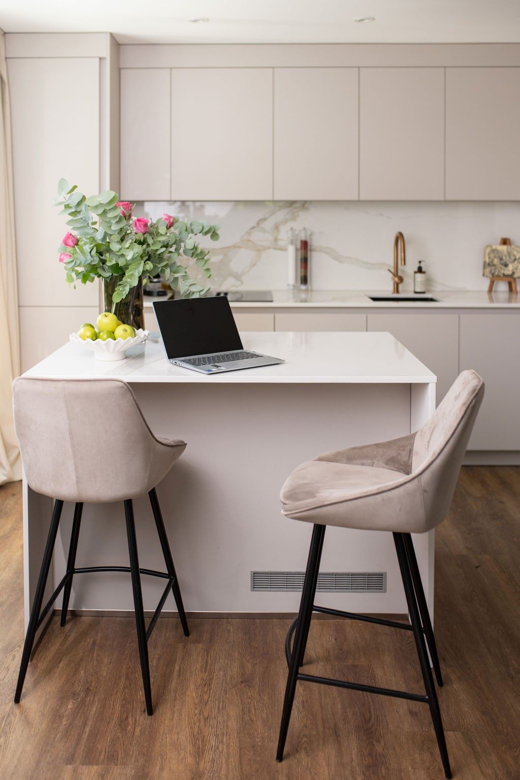A kitchen with a cooking island made of white marble, in front of it are two beige bar stools made of velour. On the cooking island there is a laptop, a bowl of apples and a magnificent bouquet of flowers.