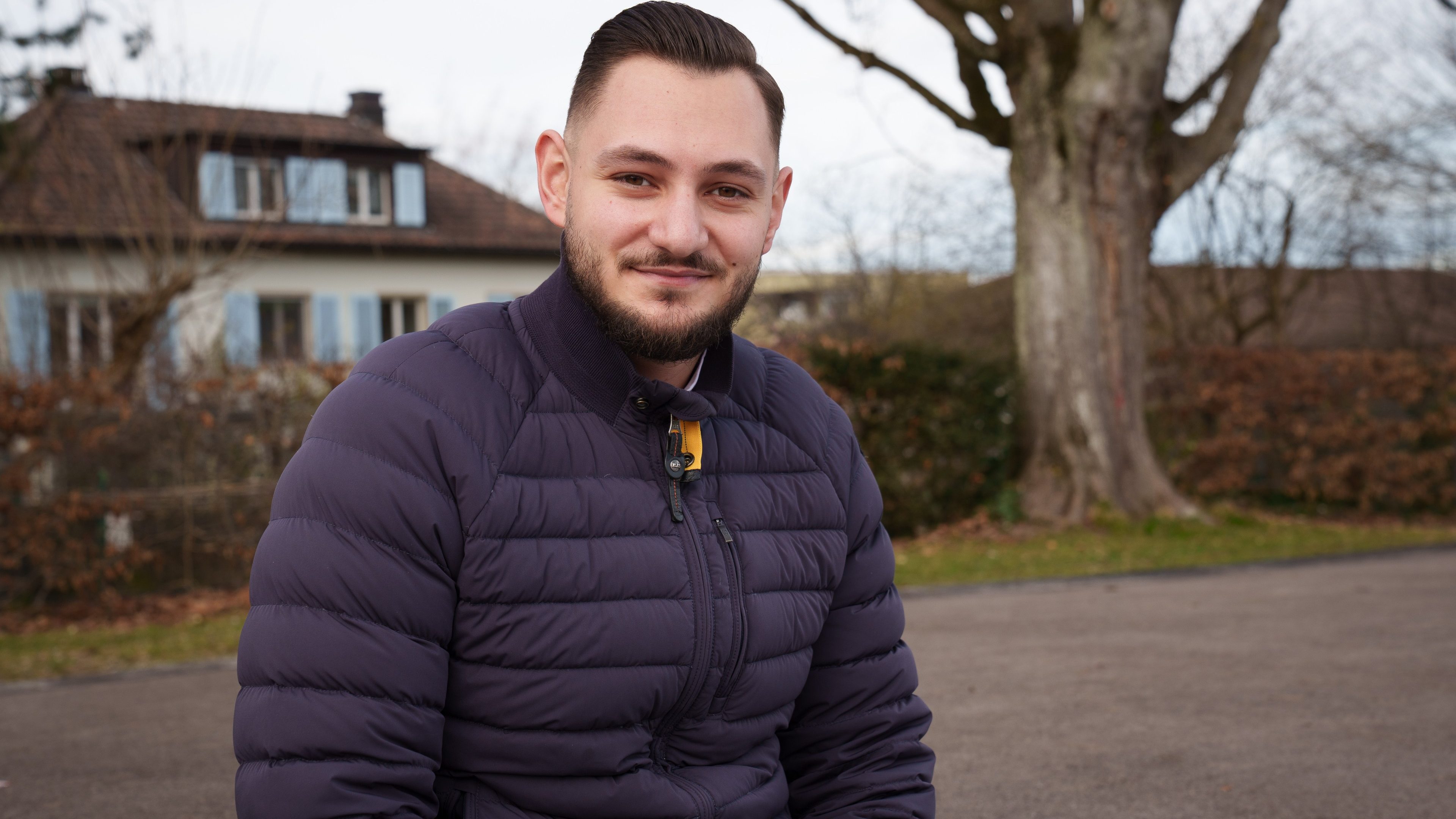 Man in down jacket stands in front of a tree and house in the background.