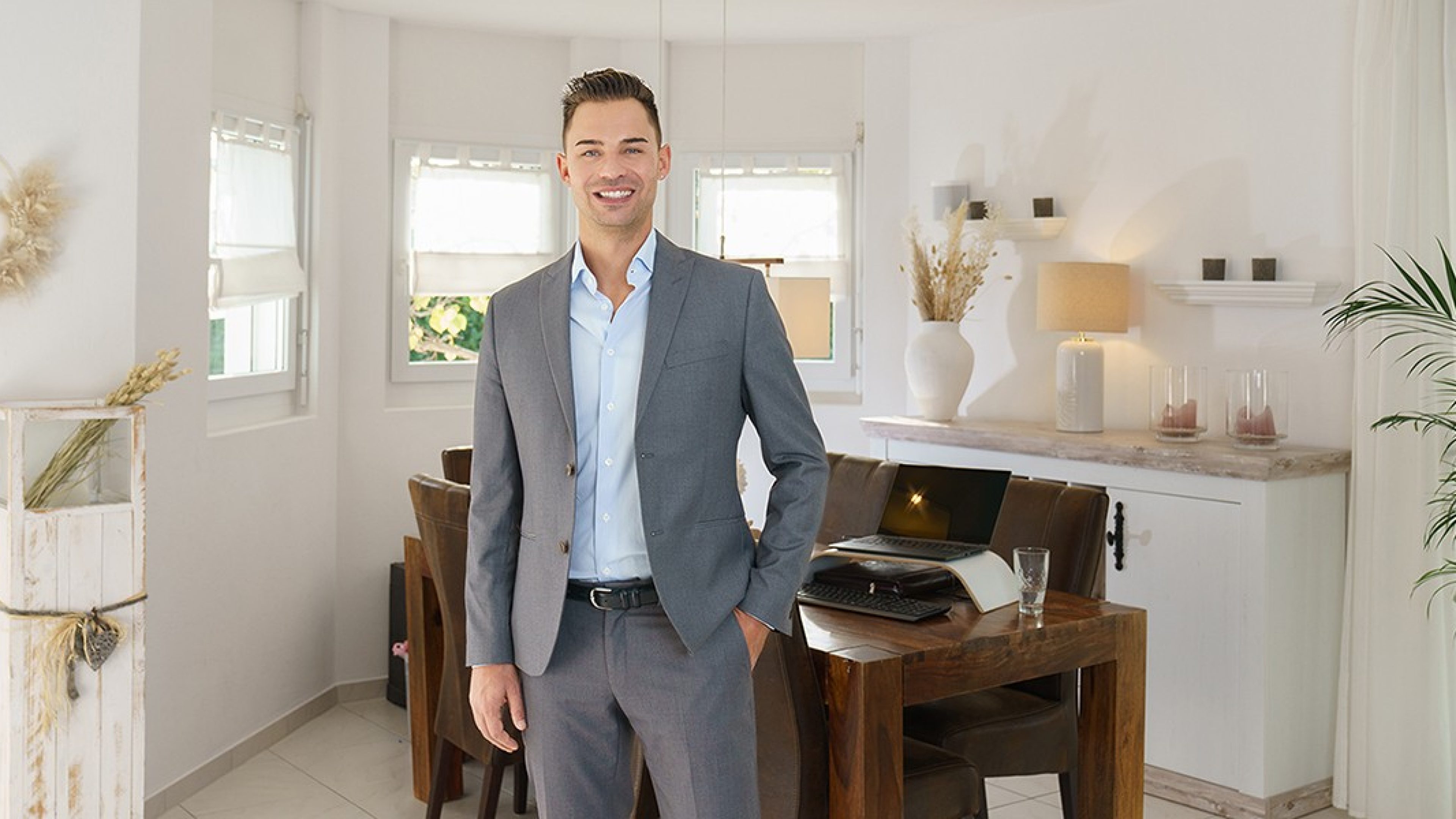 Man in business outfit stands in front of a table with a laptop and smiles at the camera.