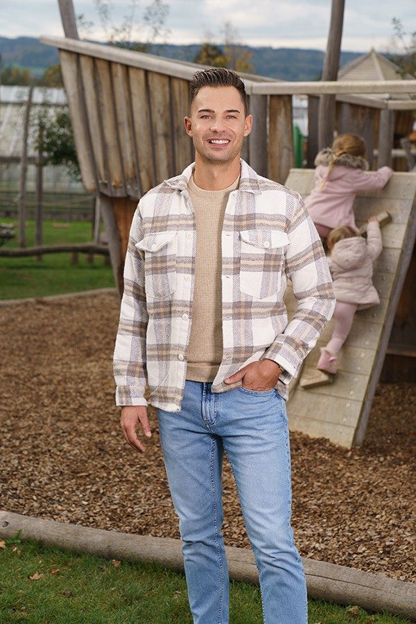 : Man stands on a playground and smiles at the camera, behind him two small children play