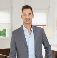Man in business outfit stands in front of a table with a laptop and smiles at the camera.