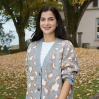A woman wearing a patterned cardigan walks towards the camera, behind her there are fallen leaves on the ground.