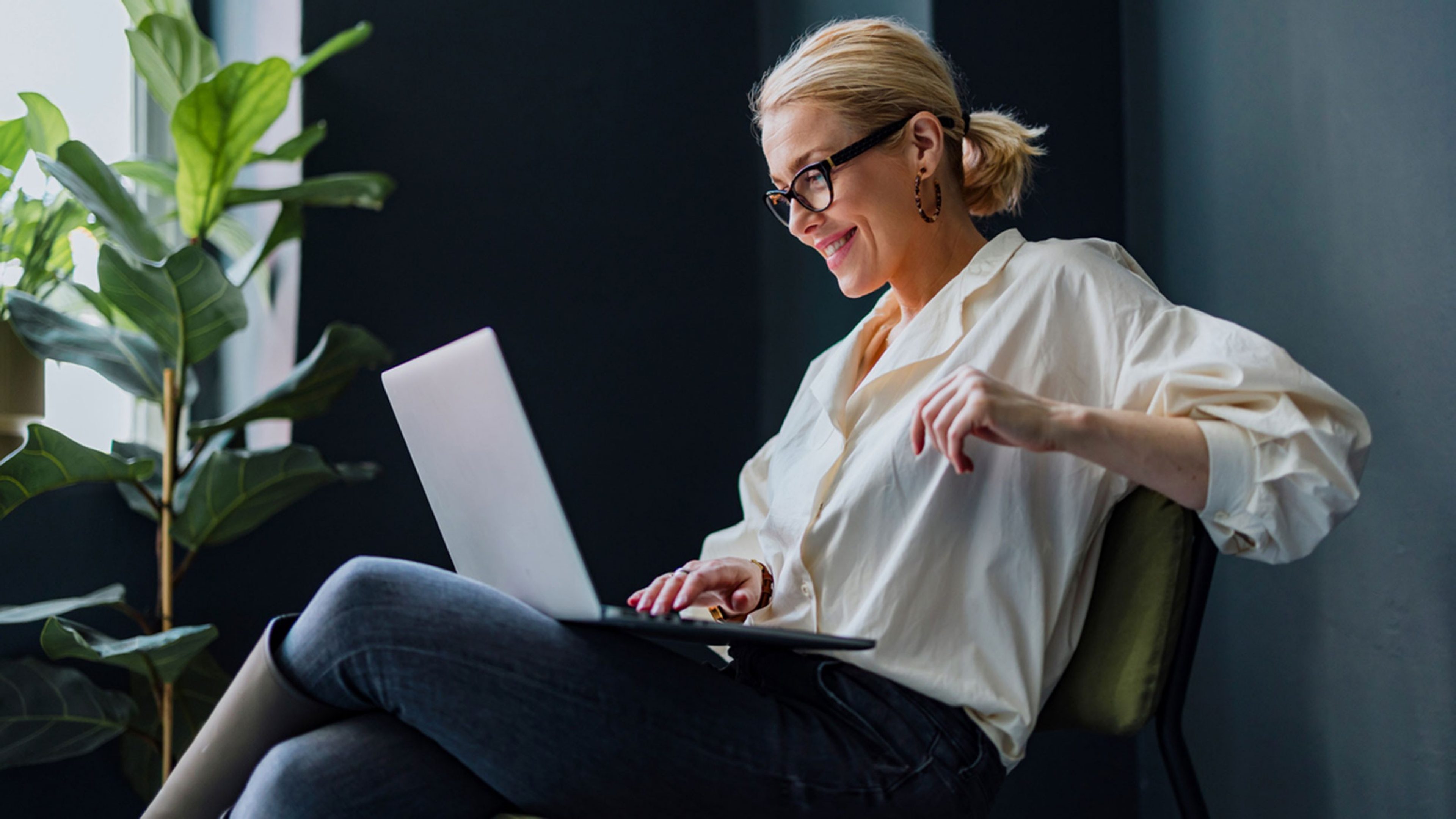A woman wearing glasses and earrings is sitting on a chair with a laptop on her knee