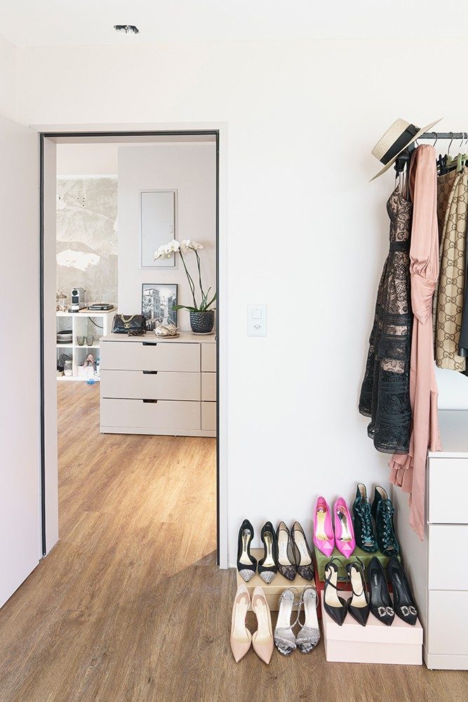 A door behind which is a chest of drawers, next to the door are shoes lined up on shoeboxes and a clothes stand with clothes.  