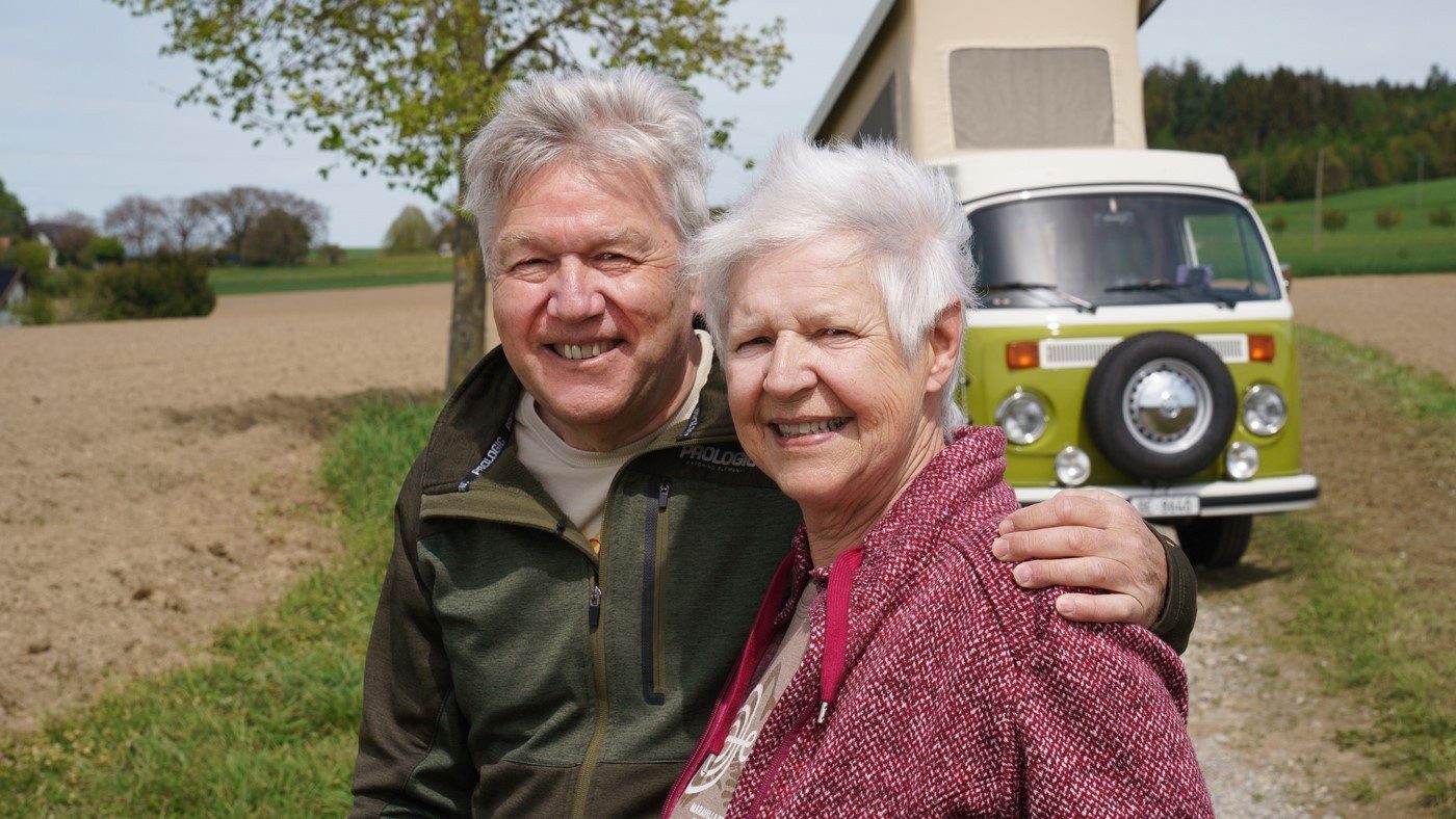 Kurt has his left arm around Monica while they look into the camera with a smile in front of their moss-green VW van. They are wearing dark green and dark red sweaters.